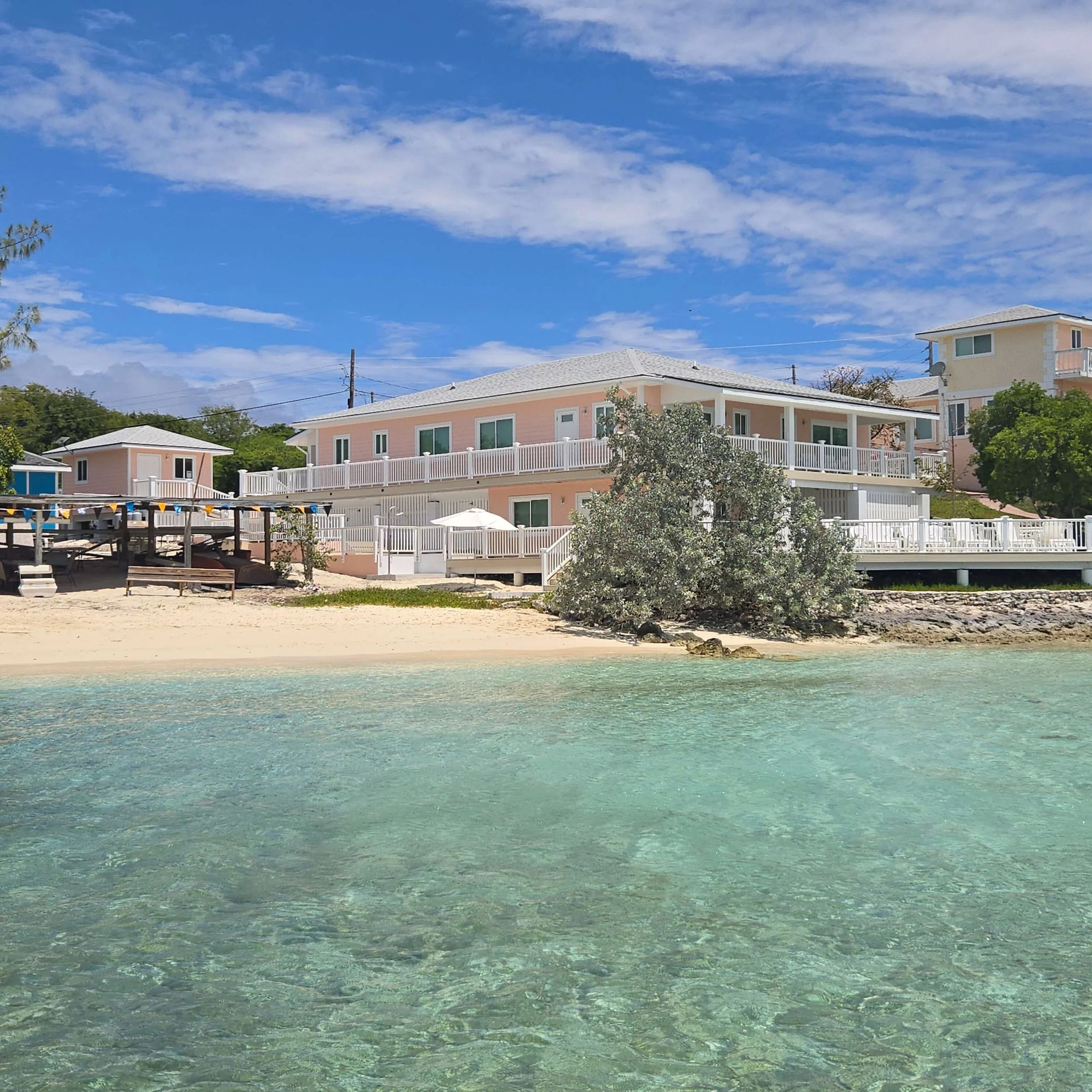 View of a beach with clear turquoise water, sandy shore, and pastel-colored houses with white fences under a partly cloudy blue sky.
