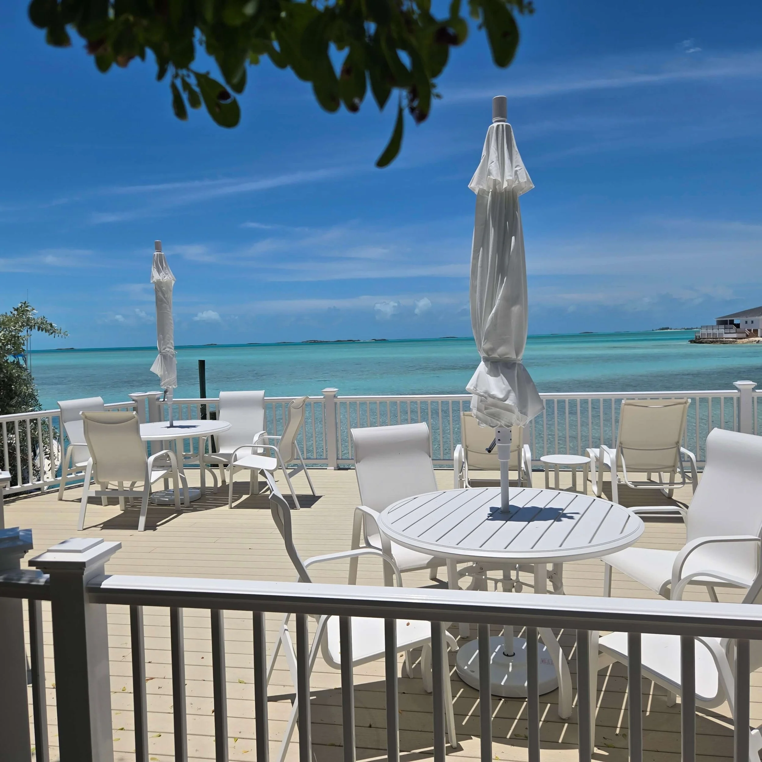 White patio furniture with closed umbrellas on the Seaside deck overlooking Staniel Cay beach and ocean under a blue sky.