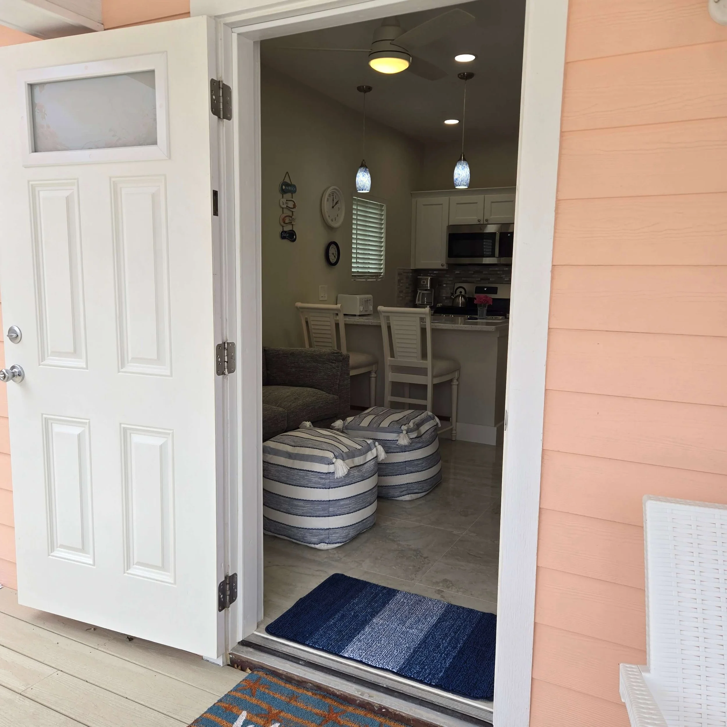 Open front door revealing a kitchen with hanging pendant lights, a wall clock, and striped ottomans in a cozy beach-themed space.