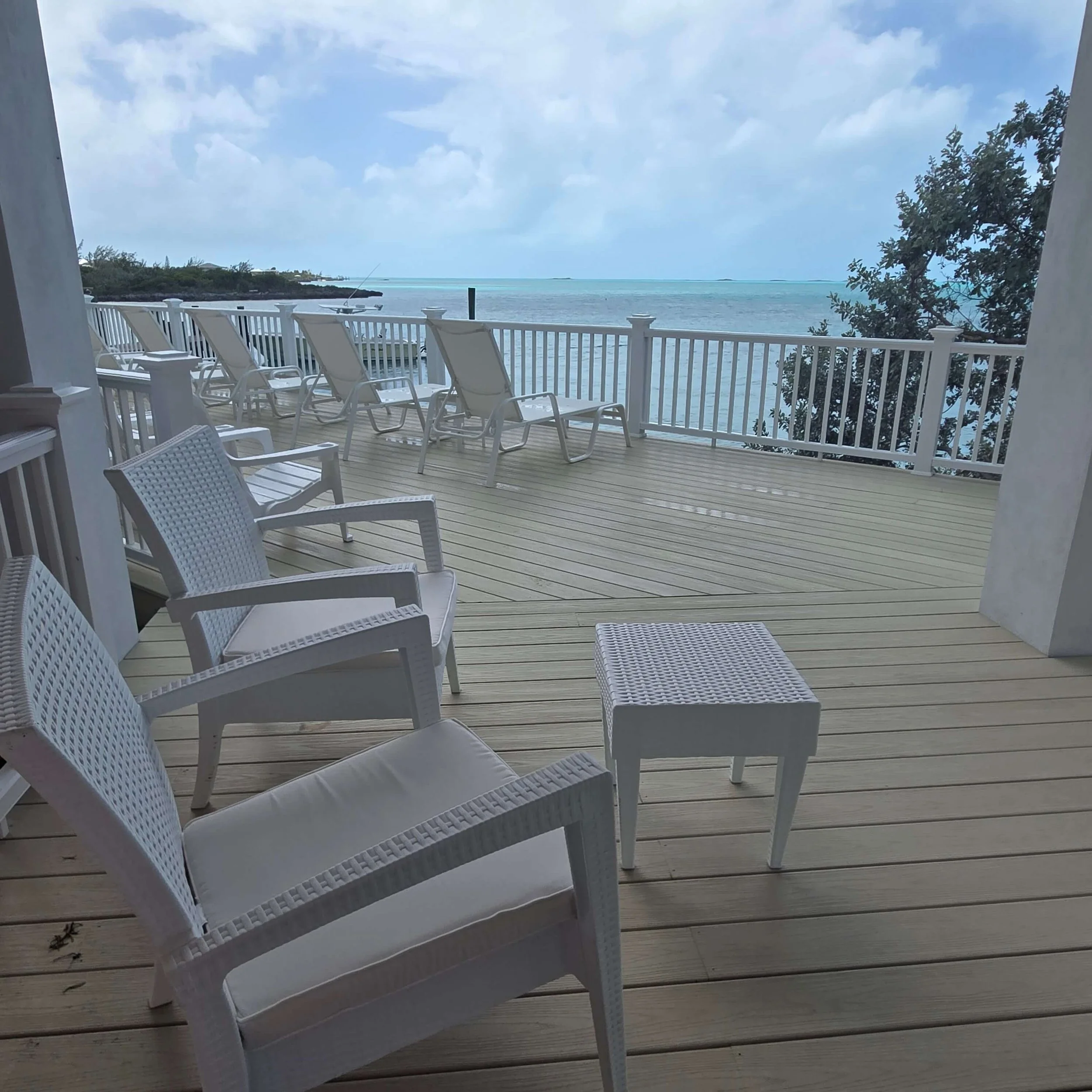 A view from a porch overlooking a body of water with a cloudy sky, white railing, and several white lounge chairs on a wooden deck.