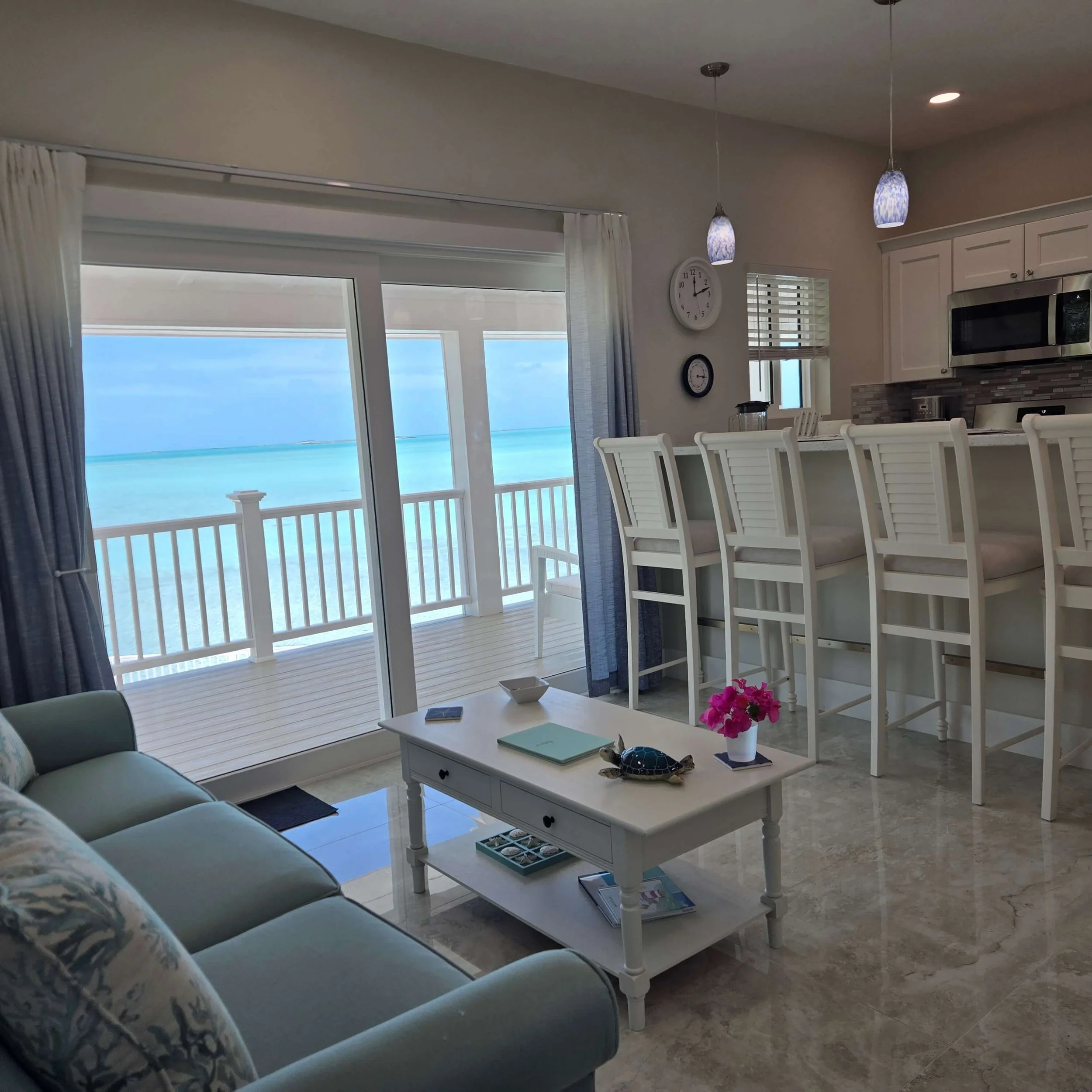 Living room with a sliding glass door looking out to an ocean view, a light blue sofa with floral pillows, a white coffee table with pink flowers, and a kitchen with white chairs, a white counter, and hanging pendant lights.