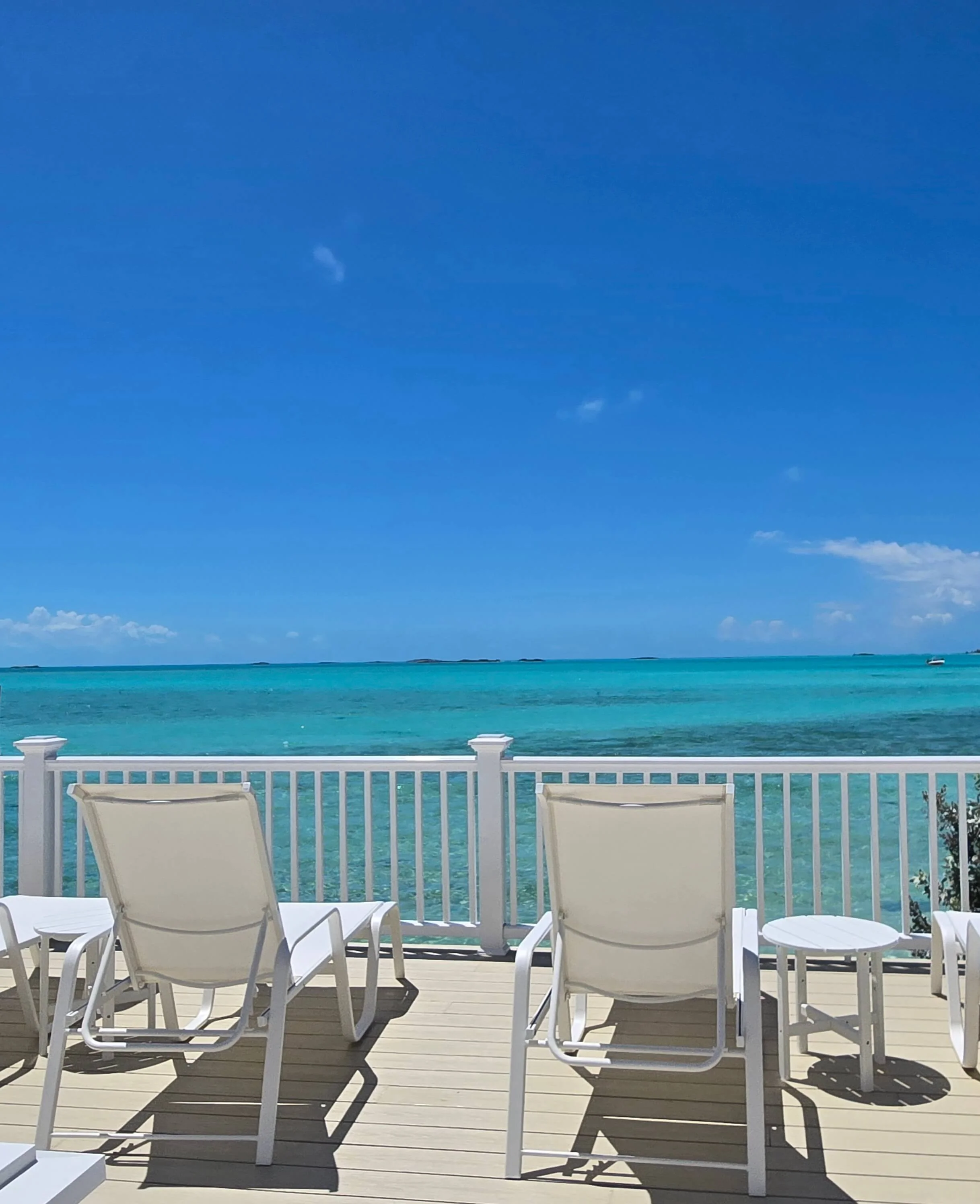 Seaside beachside patio with white chairs and tables overlooking turquoise ocean, clear blue sky, and distant small islands 