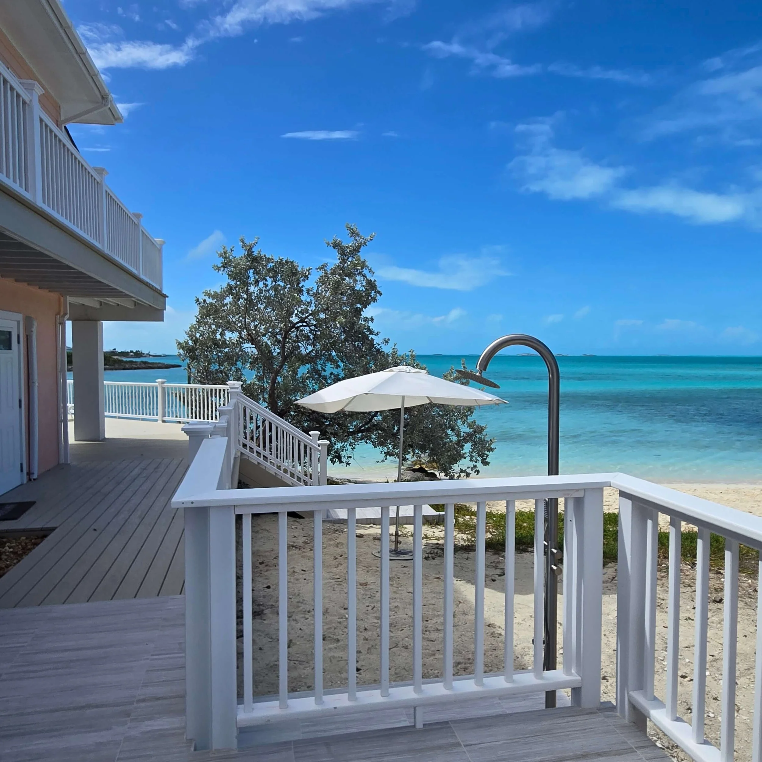View of a beachfront house with white railings and a deck, overlooking the ocean with a clear blue sky, some clouds, trees, and an umbrella.