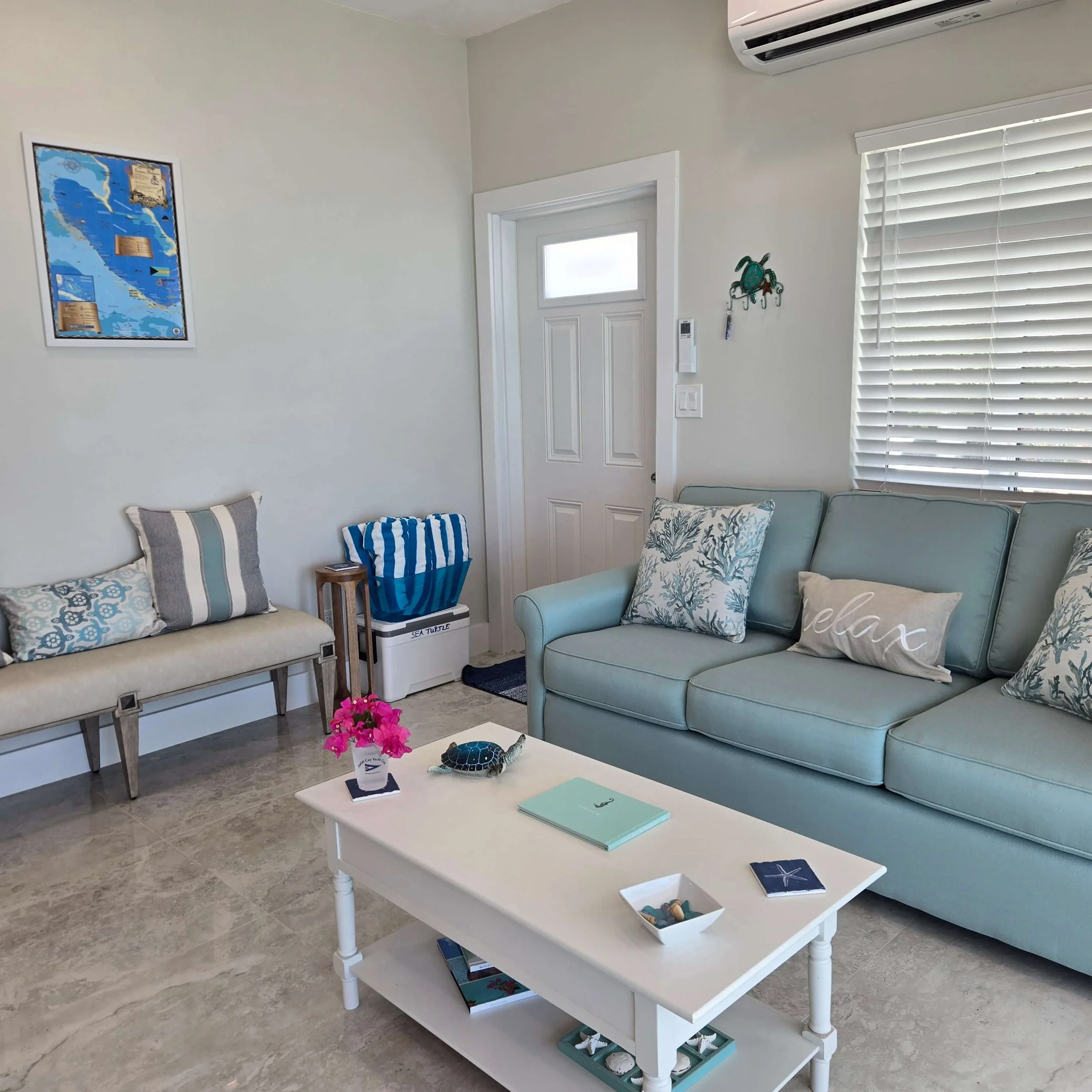 Living room with white walls, gray tiled floor, and light blue furniture including a sofa and armchair, adorned with patterned pillows. A white coffee table holds a pink flower pot, books, and a decorative turtle figurine. A wall-mounted air conditio