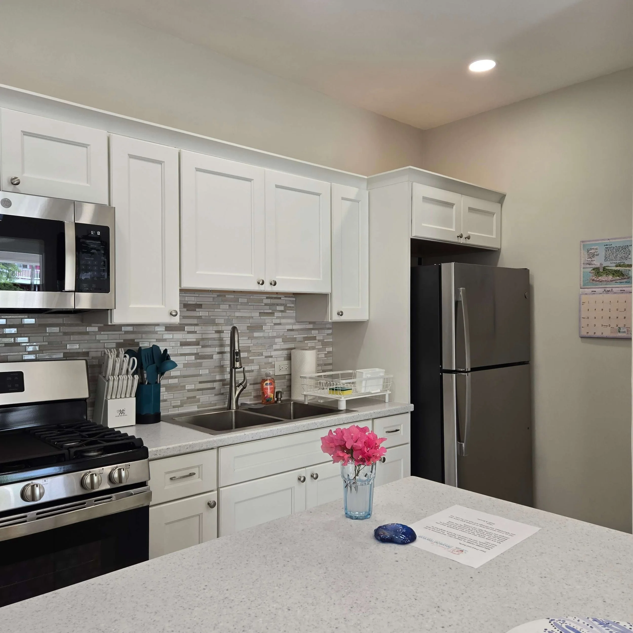 A modern kitchen with white cabinets, a stainless steel microwave, a stove, a double sink, and a refrigerator. There is a blue glass vase with pink flowers on the kitchen island and a paper with text. The backsplash has a gray and white mosaic tile p