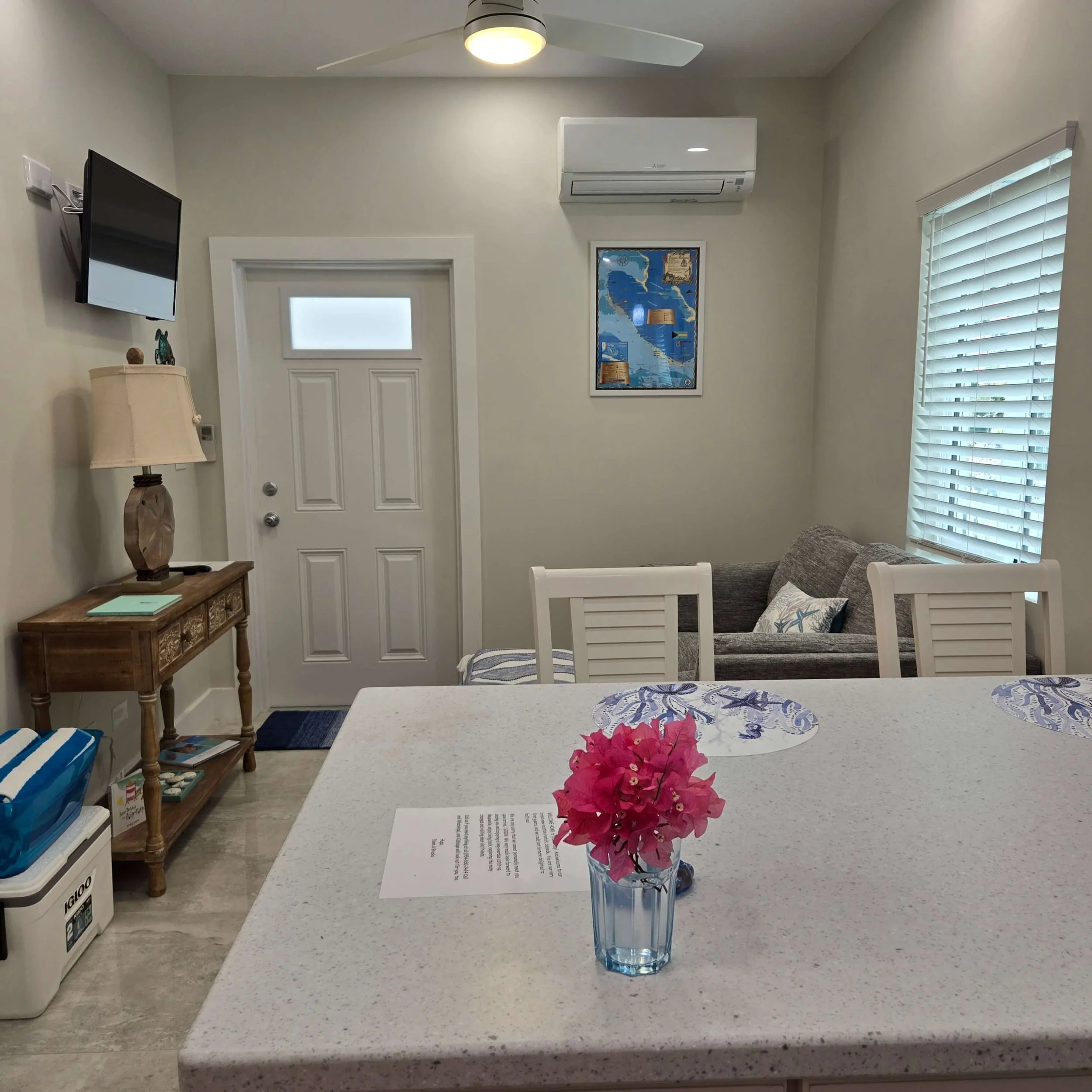 A kitchen with a white countertop, pink flowers in a clear blue vase, a white dining chair, behind which there is a gray couch, and a wall with a map and windows with white blinds.