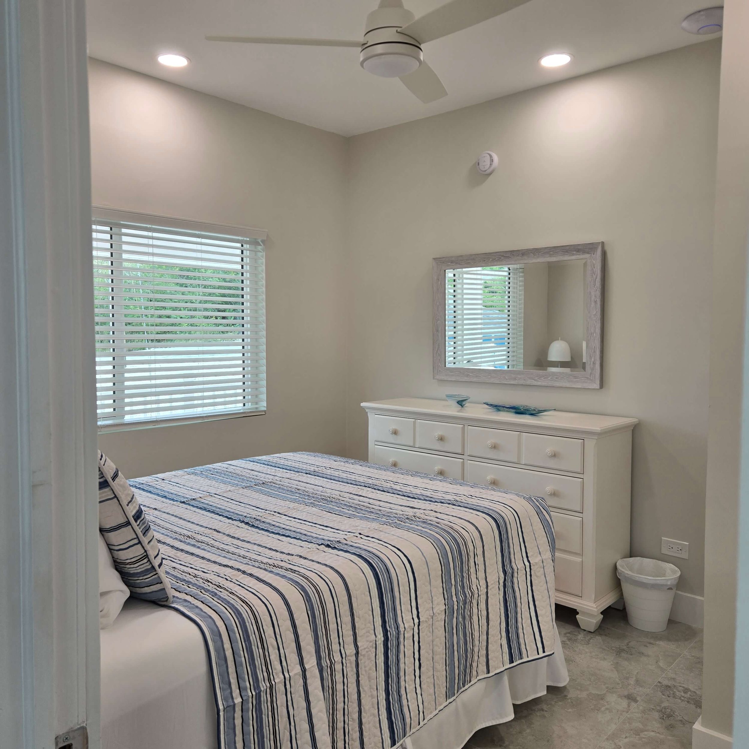 A bedroom with two windows with white blinds, a white dresser with three rows of drawers, a large mirror framed in light wood hanging on the wall, a striped bedspread in shades of blue, white, and beige, and a corner trash can and a small white table