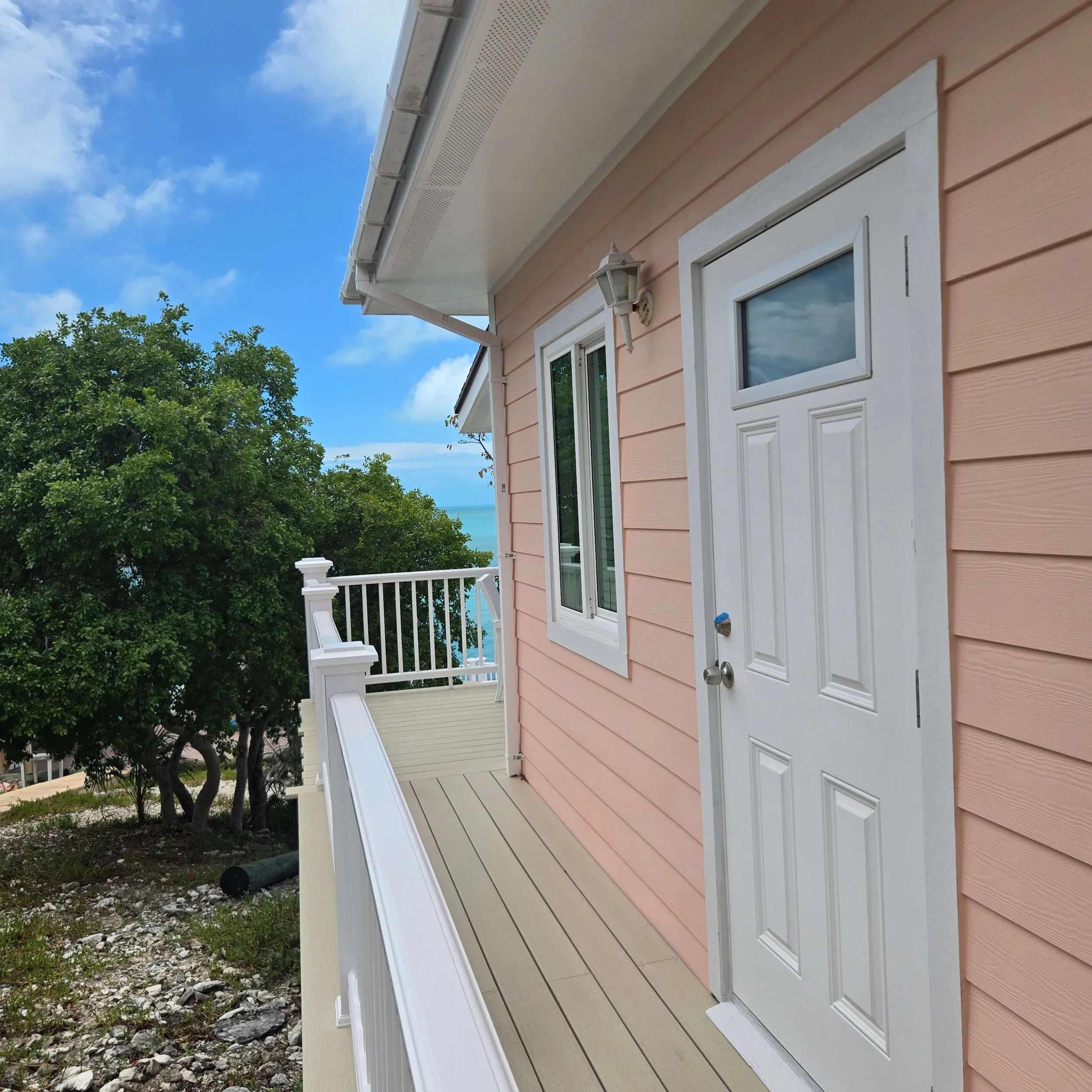 The image shows the exterior of a house painted in pink with white details, a porch with white railing, a door with a window, and a wall-mounted lantern light. There are trees and a partly cloudy blue sky in the background.