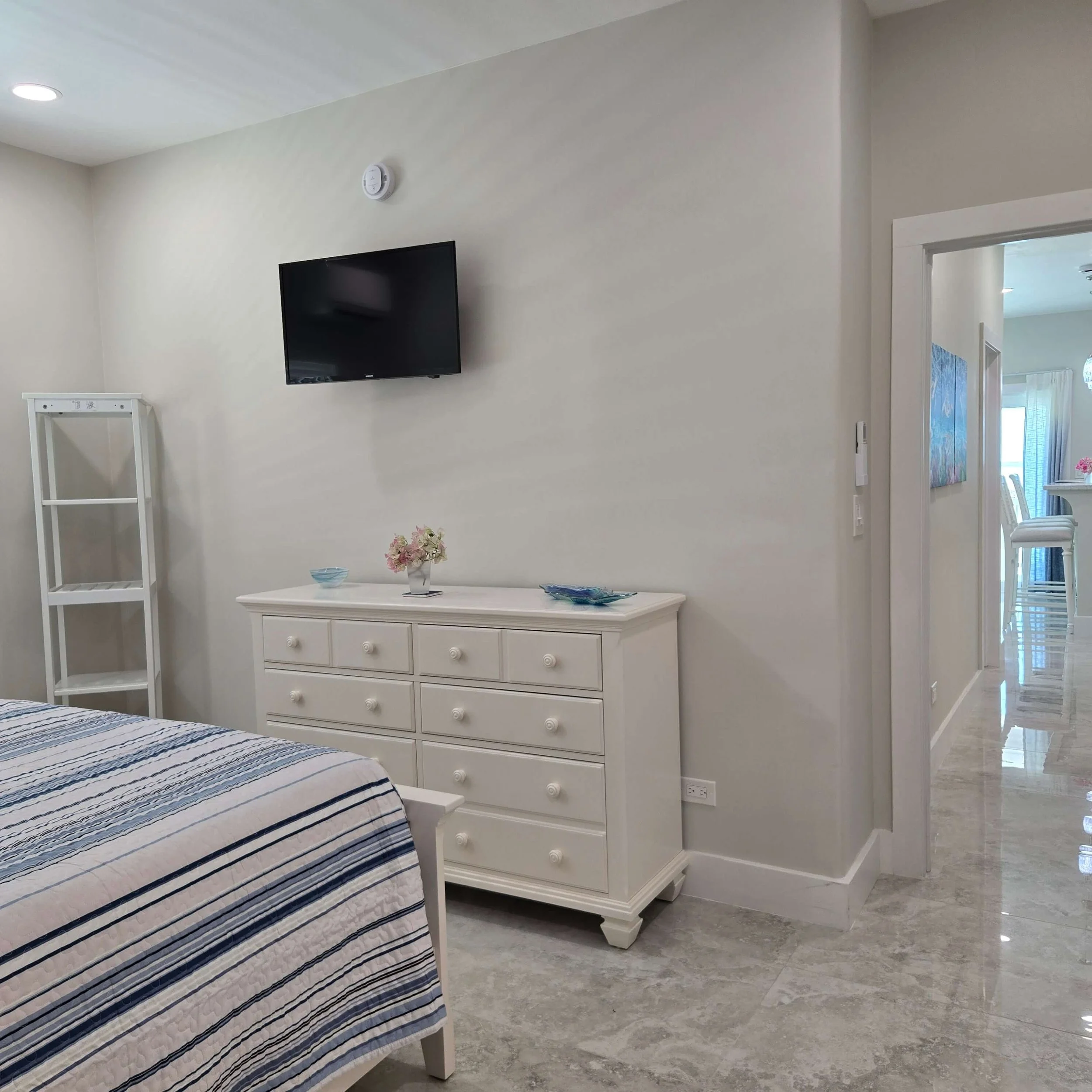 A bedroom with a white dresser, a television mounted on the wall, a part of a bed with a striped blanket, candles, and flowers on the dresser, and a doorway leading to another room.