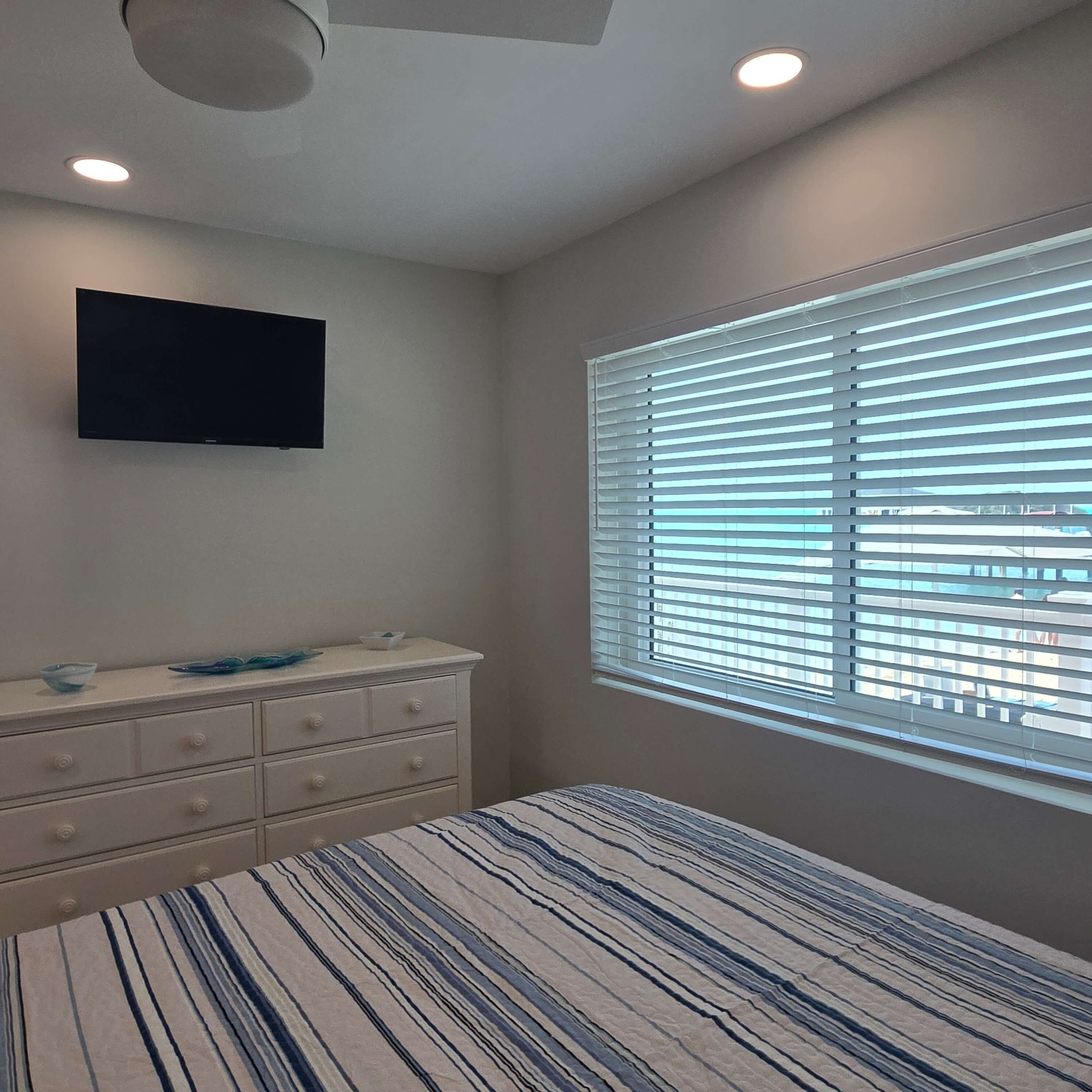A bedroom corner with a striped rug on the floor, a dresser with bowls on top, a wall-mounted flat screen TV, a window with white blinds, and part of a bed with striped bedding.
