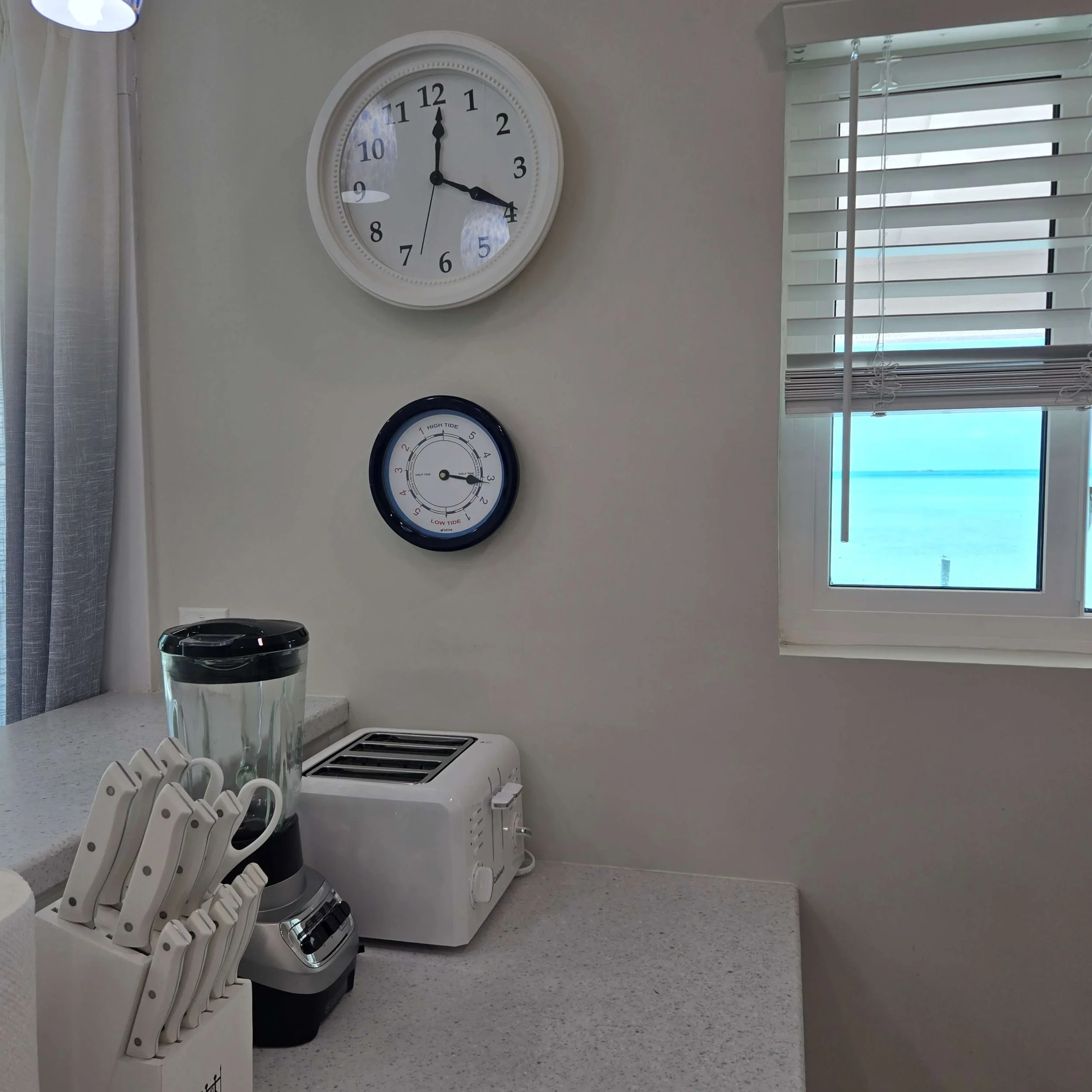 Kitchen with a wall clock showing 3:00, a window with blinds, a countertop, a set of knives in a knife block, a microwave oven, and a trash can.