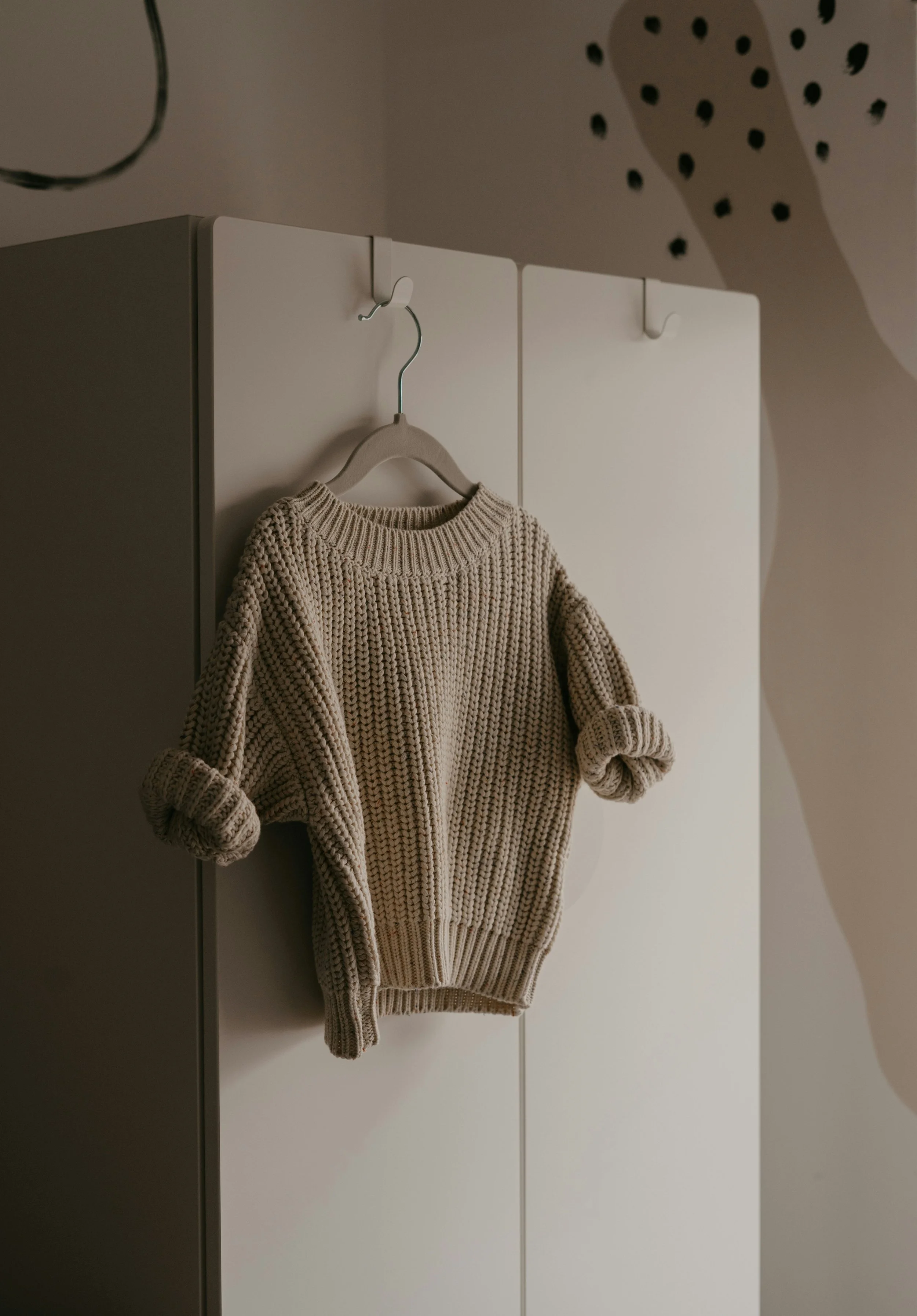 A beige knitted sweater hangs on a white hanger, attached to a hook on a closed white wardrobe door.
