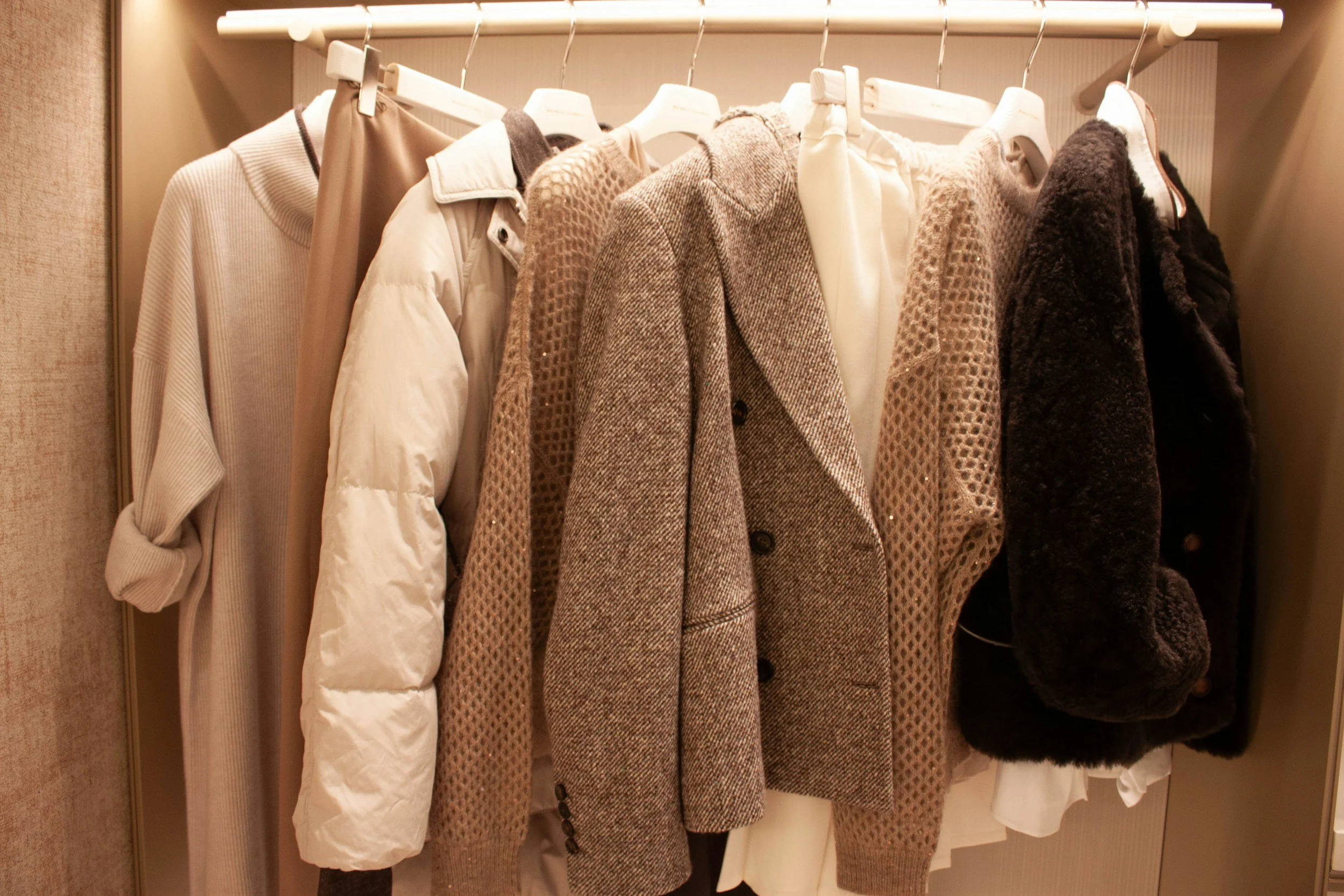 A row of coats and jackets hanging on a clothing rack. The garments are in various shades of beige, brown, and black, with some textured and patterned fabrics.