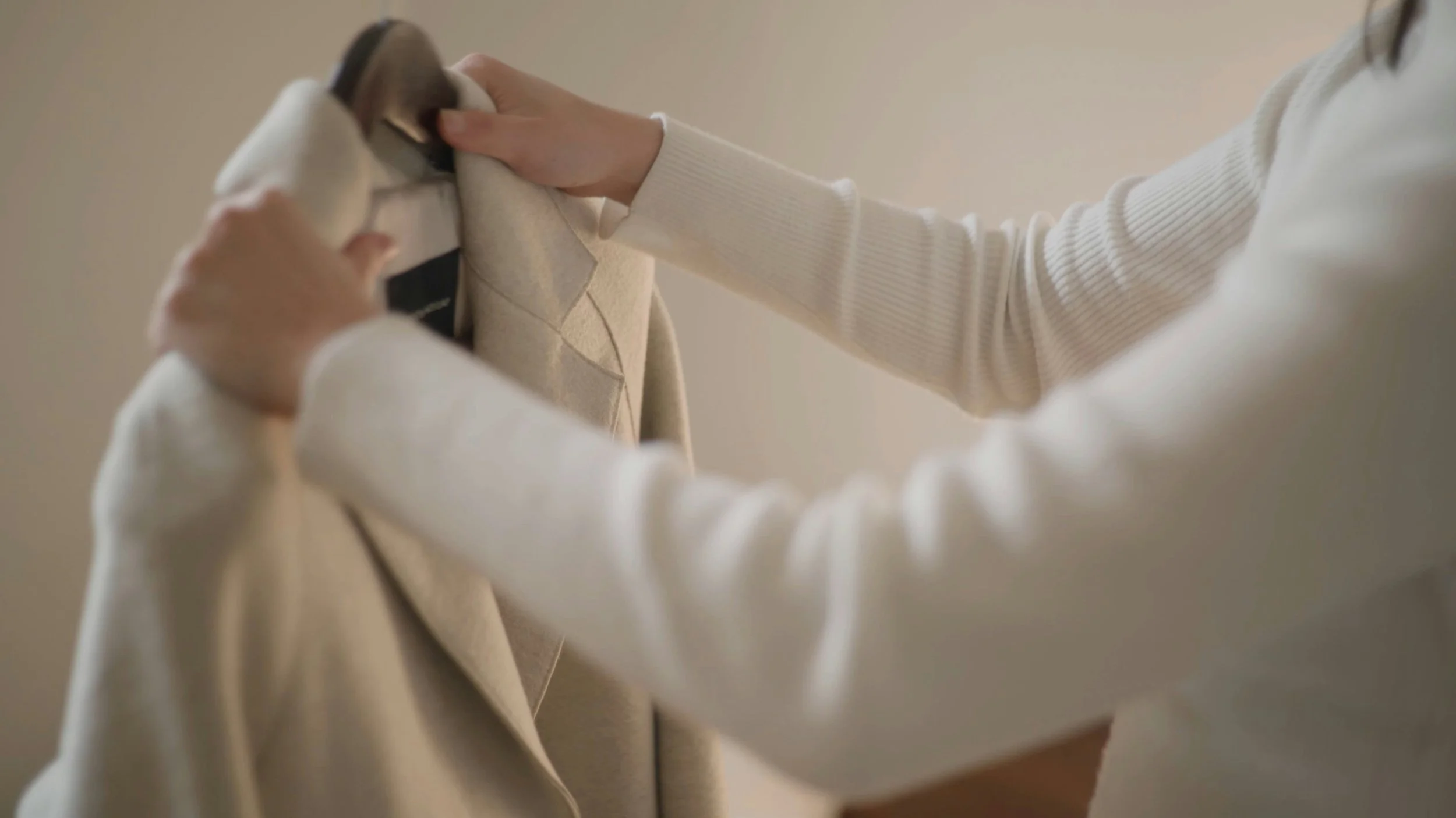 A person is hanging a beige coat on a wall hook.