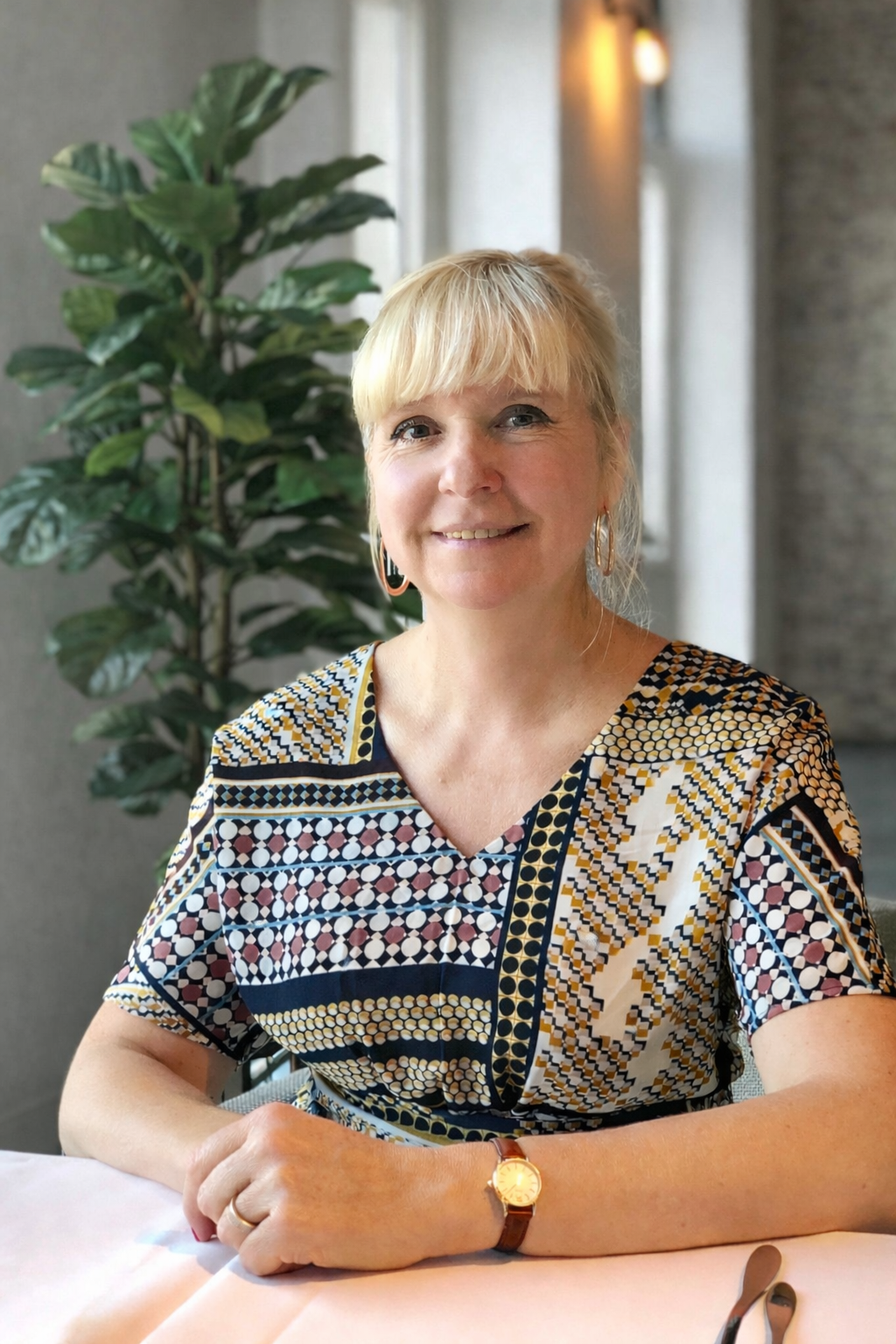 Een vrouw met blonde haar en een kleurrijk geometrisch patroon shirt zit aan een tafel met een plant op de achtergrond.