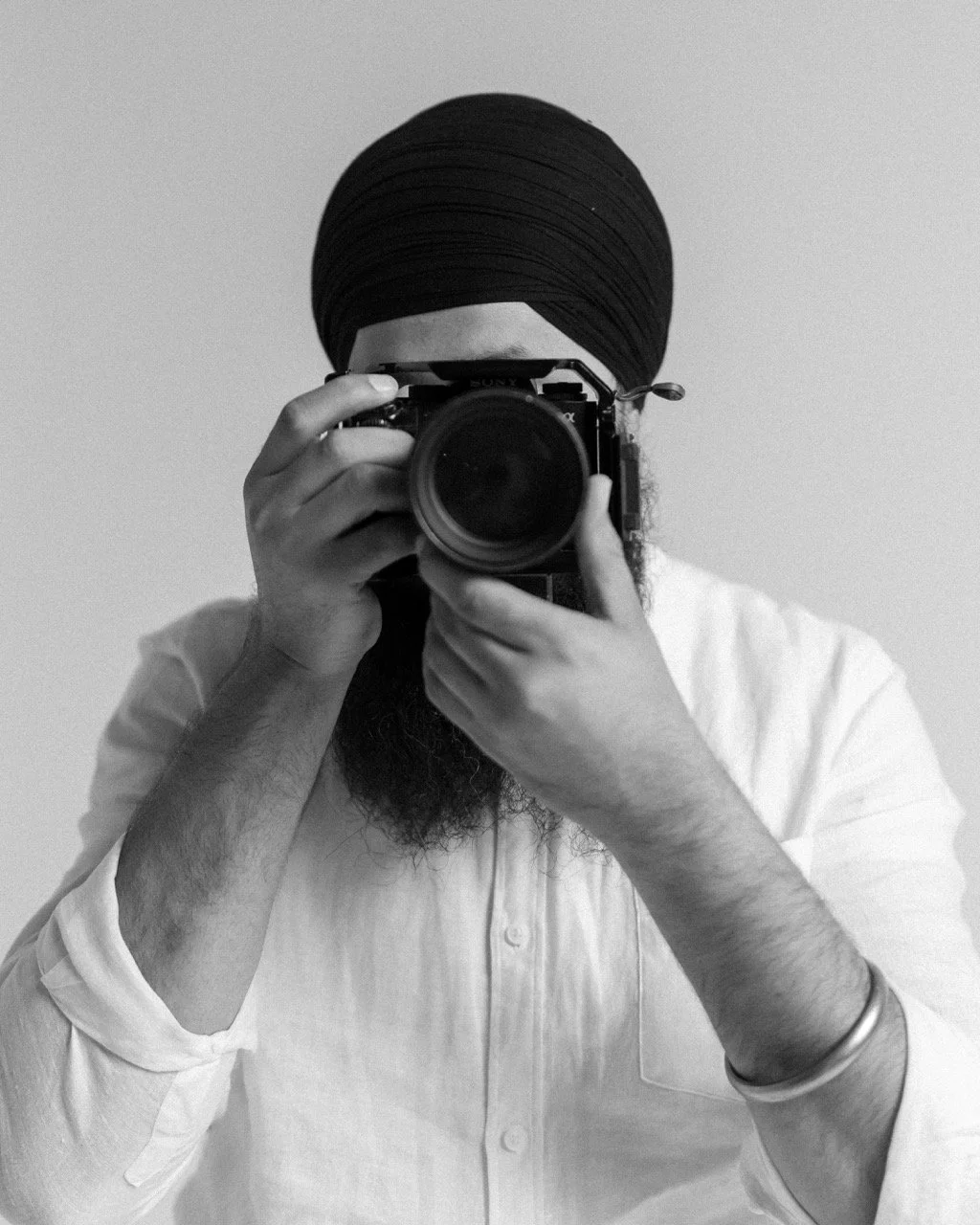 Person with a beard and turban taking a photo with a camera in front of a plain wall.