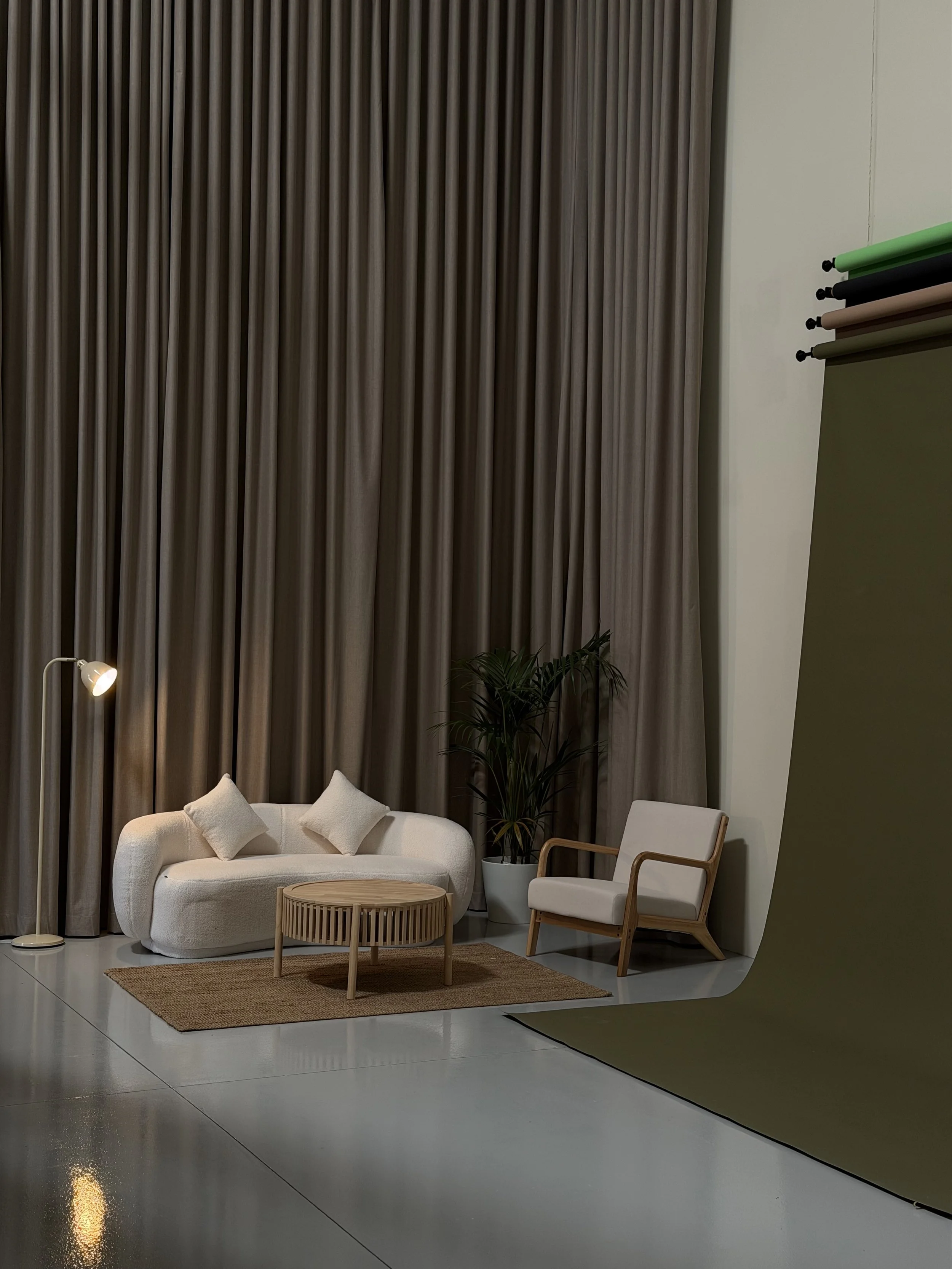 A cozy living room corner with a white curved sofa, a small round wooden coffee table, a beige armchair with wooden arms, a tall green plant in a white pot, a nested rug, beige curtains, a floor lamp, and a green photography backdrop roll on a stand.