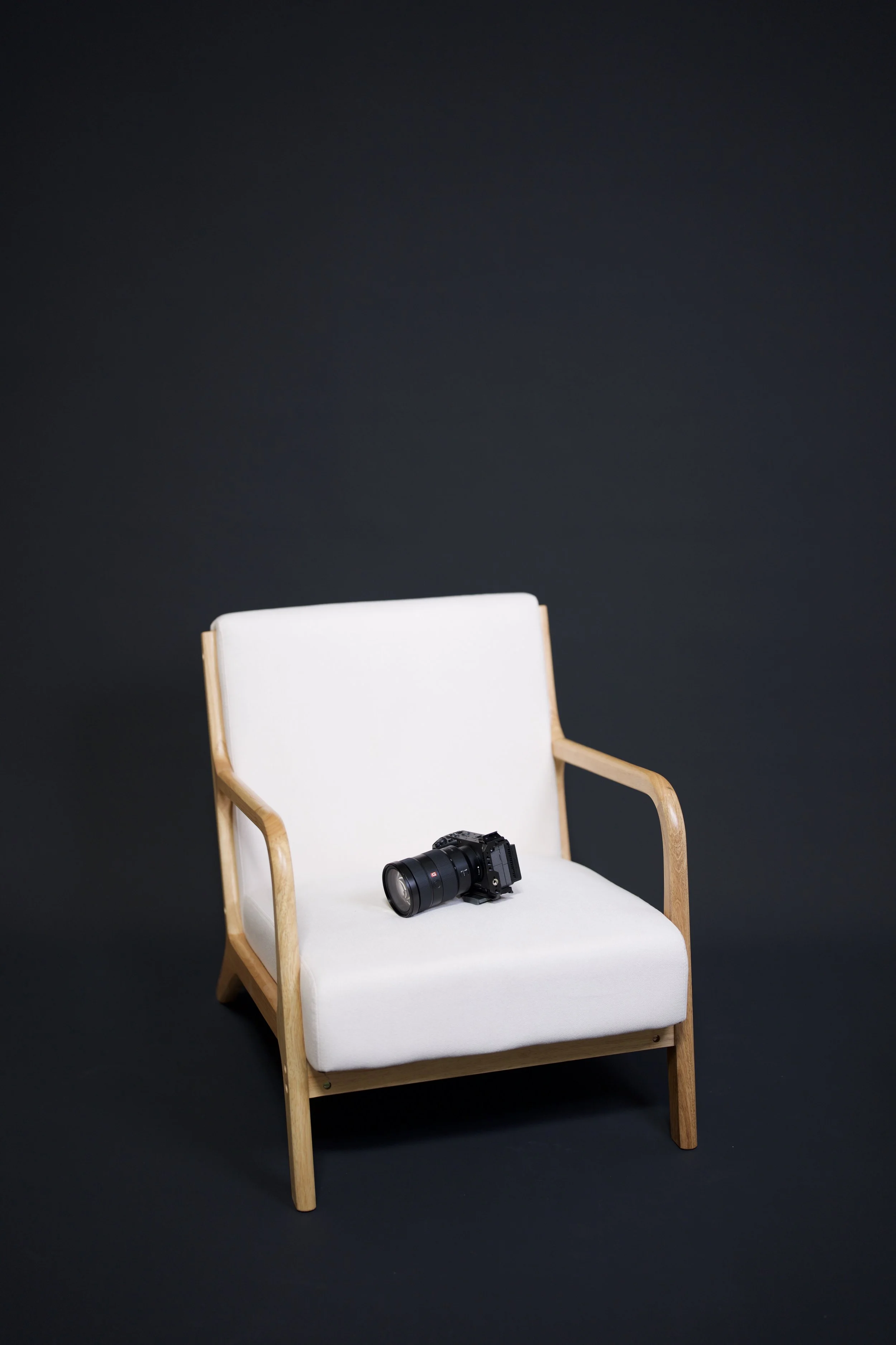 A white chair with wooden arms and legs sits against a black background. A camera is placed on the seat of the chair.
