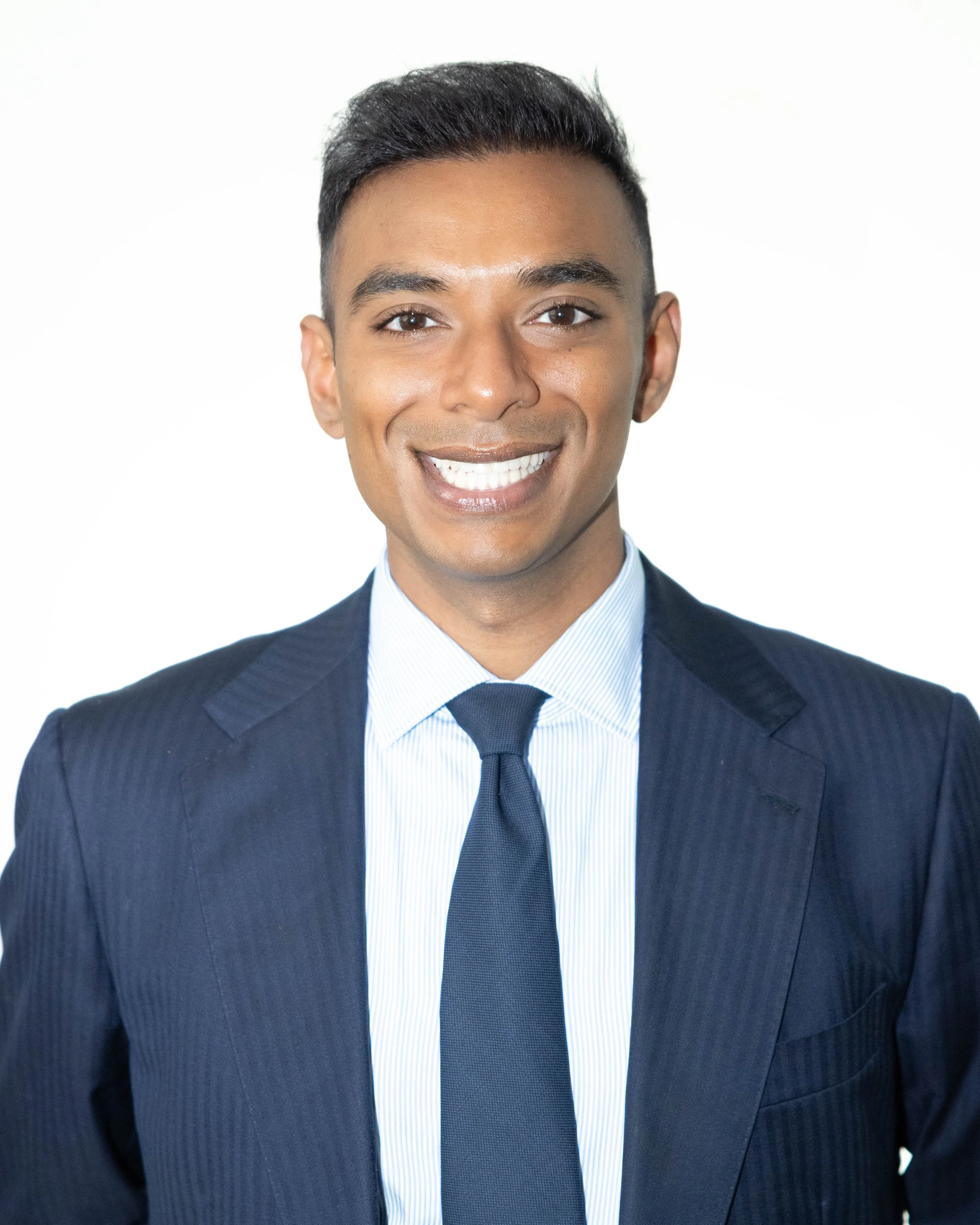 A young man with dark hair, wearing a navy blue suit, light blue shirt, and a darker blue tie, smiling against a plain white background.