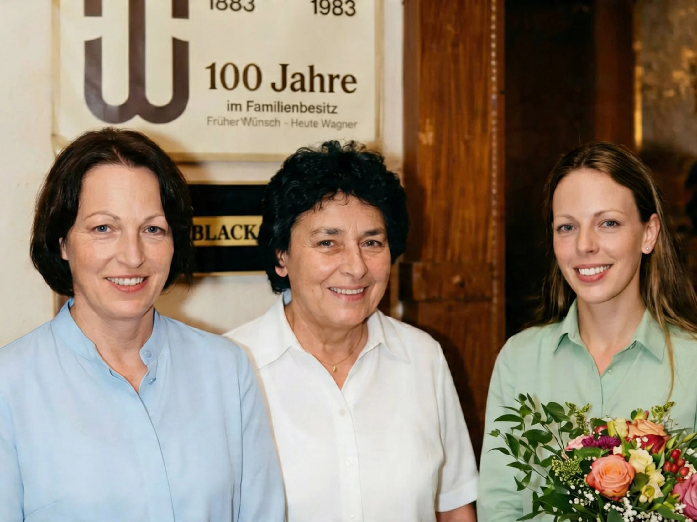 Drei Frauen stehen nebeneinander, lächeln, mit Blumenstrauß in der Hand. Im Hintergrund ein Holzregal und eine Tafel mit Jahreszahlen und Text in Deutsch.