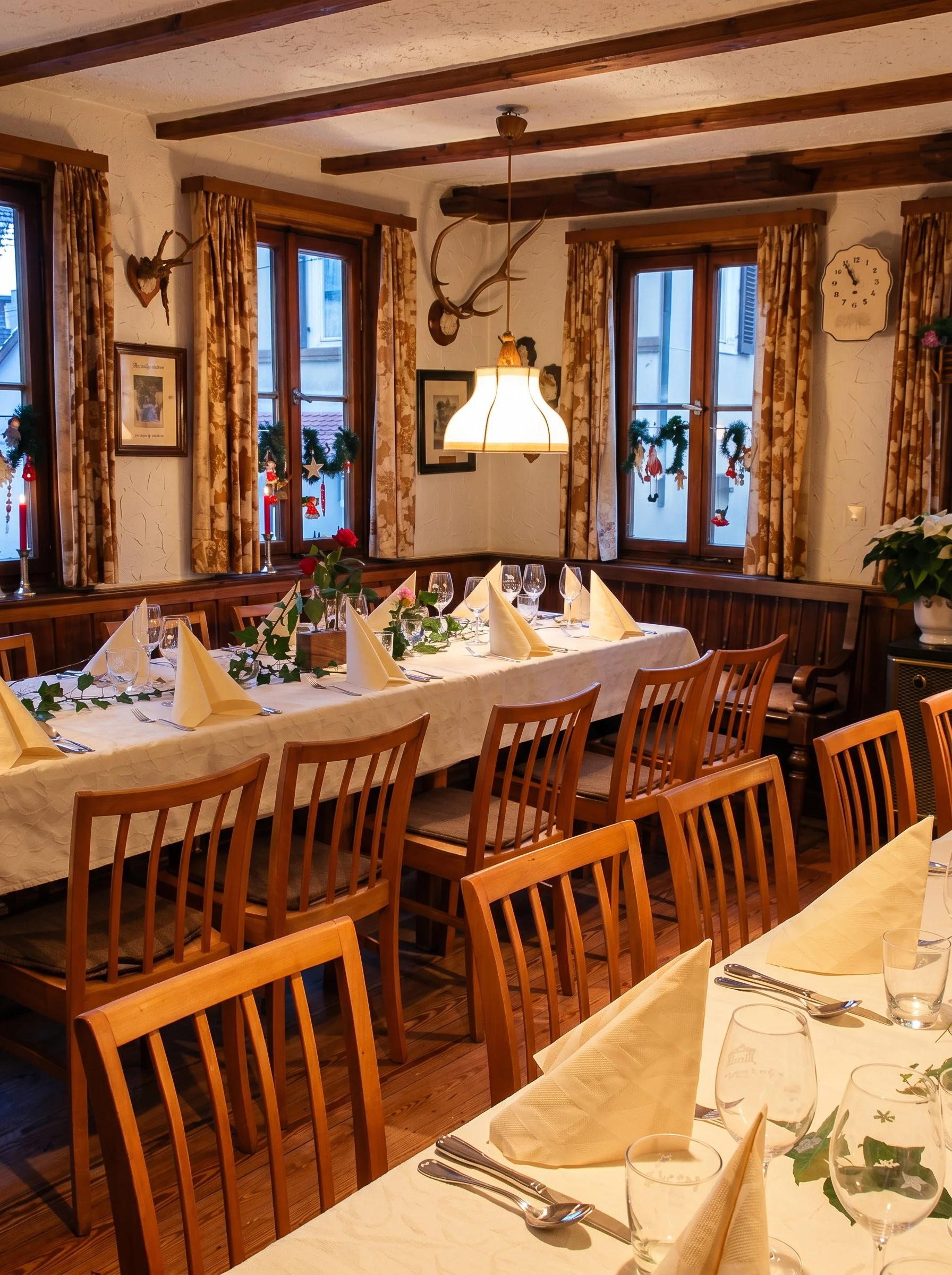 Ein festlich gedeckter Esstisch in einem rustikalen Raum mit Holzdecken, Fenster mit Gardinen, Dekorationen und Wein-gläsern.
