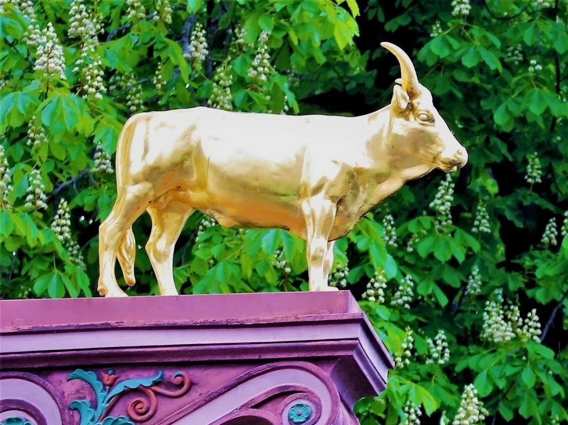 Goldene Statuette eines Rindes auf einer kunstvoll verzierten Basis vor grünem Laub.