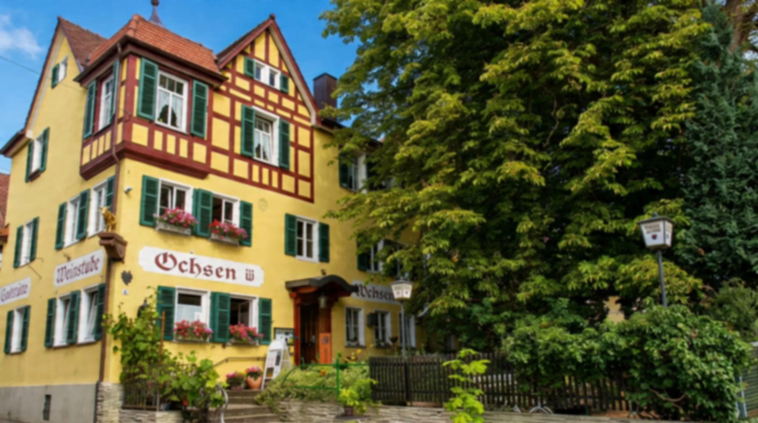 Ein bunt gestrichenes, mehrstöckiges Haus mit grünen Fensterläden und Blumen vor den Fenstern, umgeben von Bäumen, im deutschen Dorfstil.
