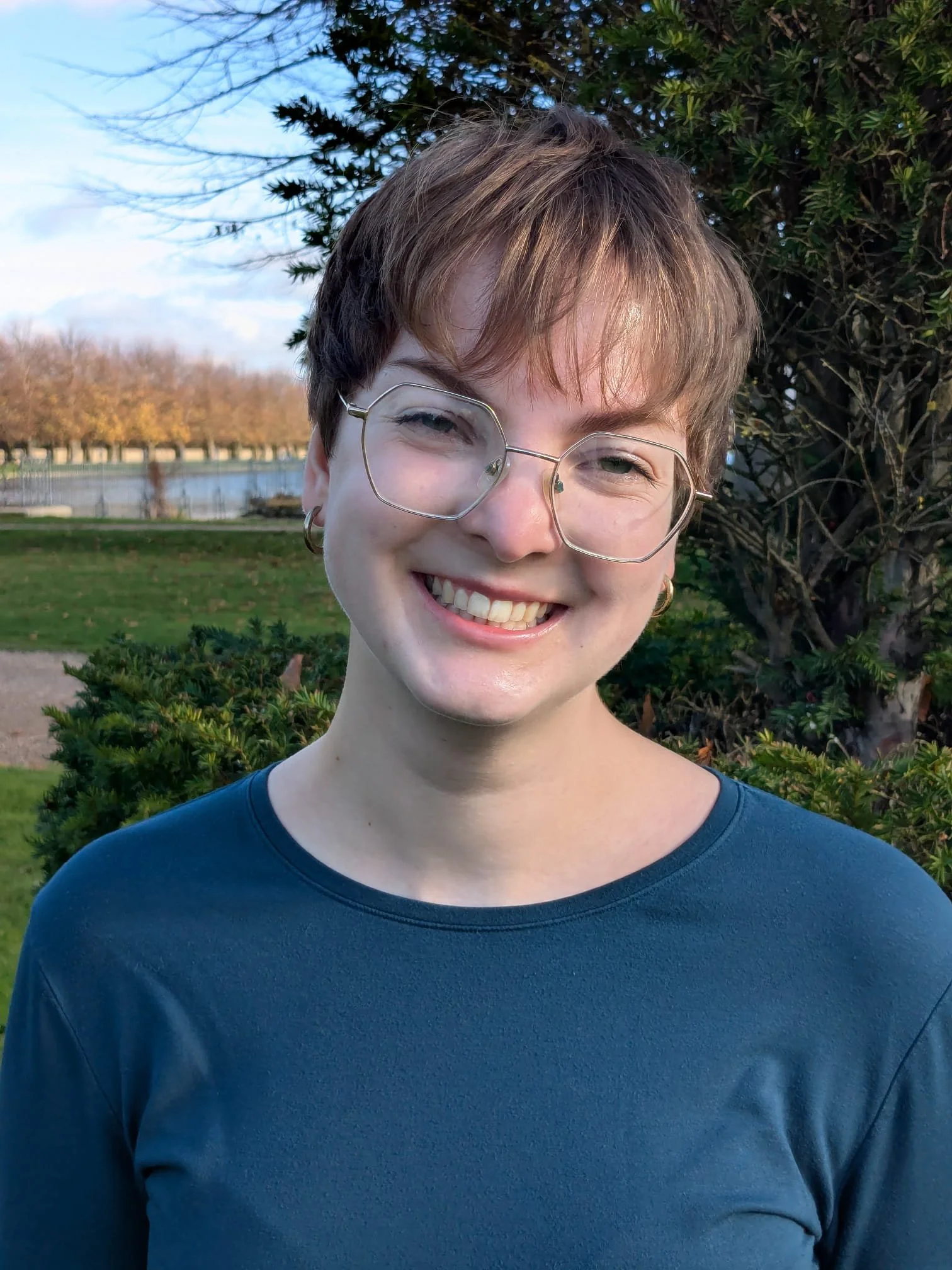 Martha is smiling. She is wearing hexagon shaped silver glasses, has short brown hair and is wearing a teal coloured top. She is standing in a sunny park.