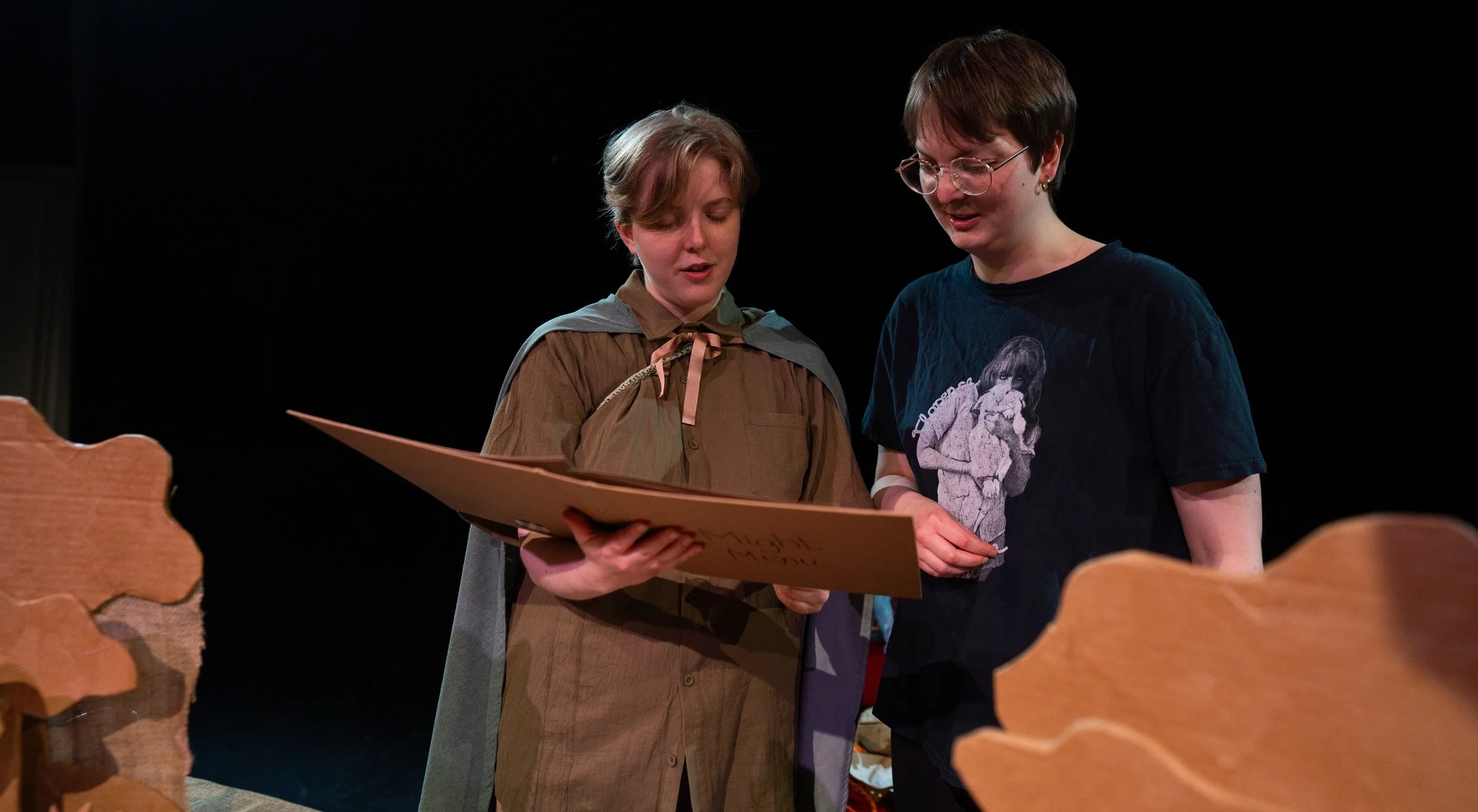 Martha and Ellie, a collaborator, look through an access prop ahead of a sharing. Ellie is in a greenn shirt and cloak, Martha is wearing glasses and is wearing a black t shirt.