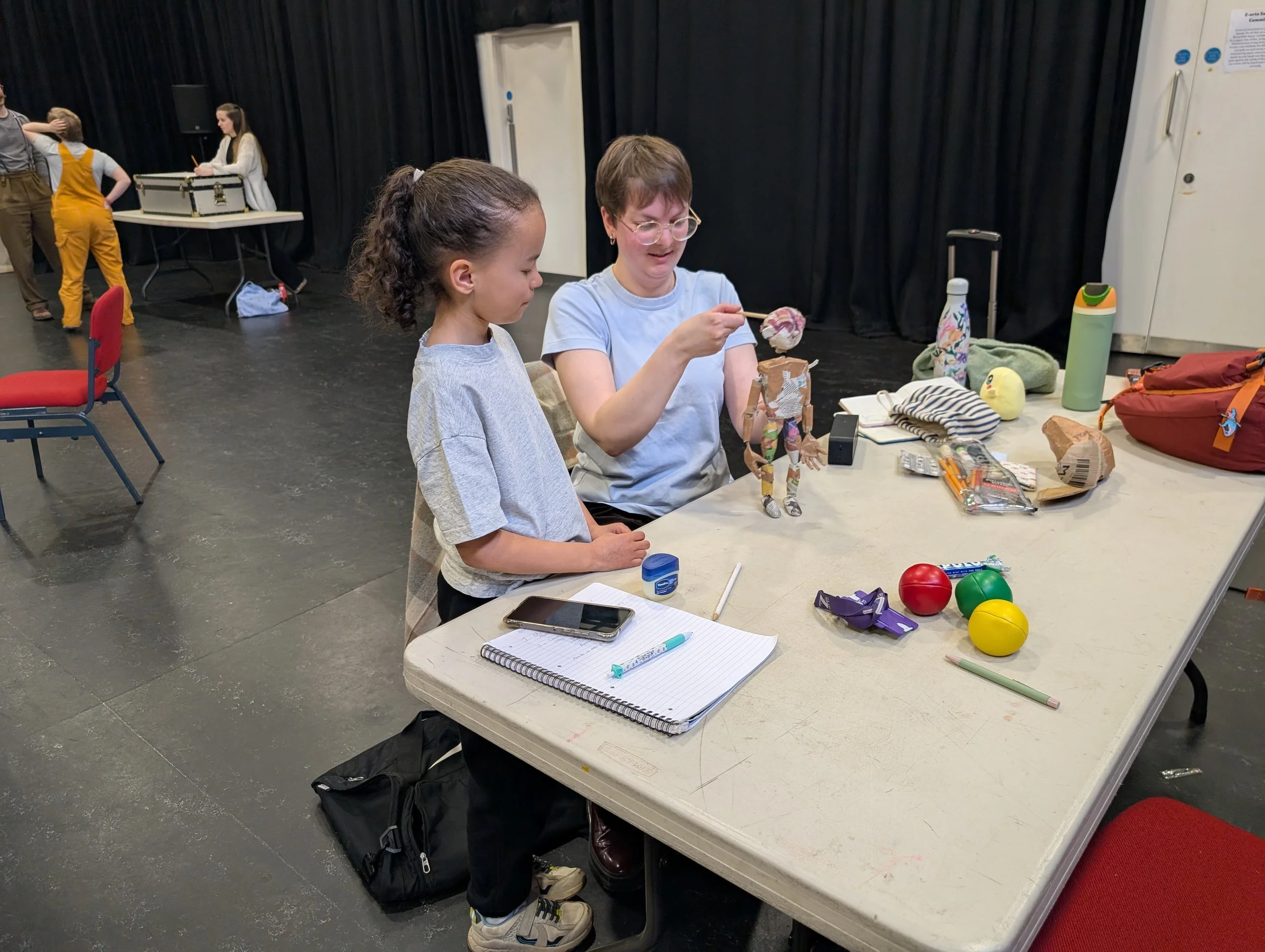 A woman and a young girl are working on a paper-mâché puppet at a table, with scattered art supplies, notebooks, a smartphone, and a water bottle in a room with black curtains and other children in the background.