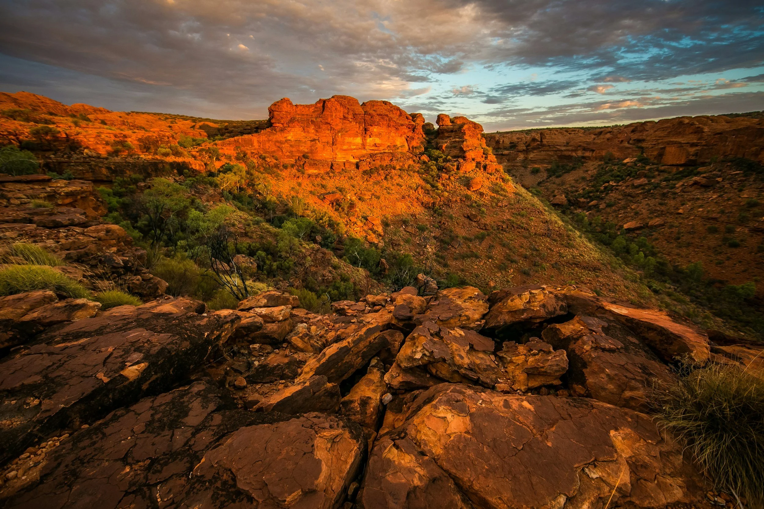 Sunset over a rocky canyon with orange and red cliffs and scattered green vegetation.