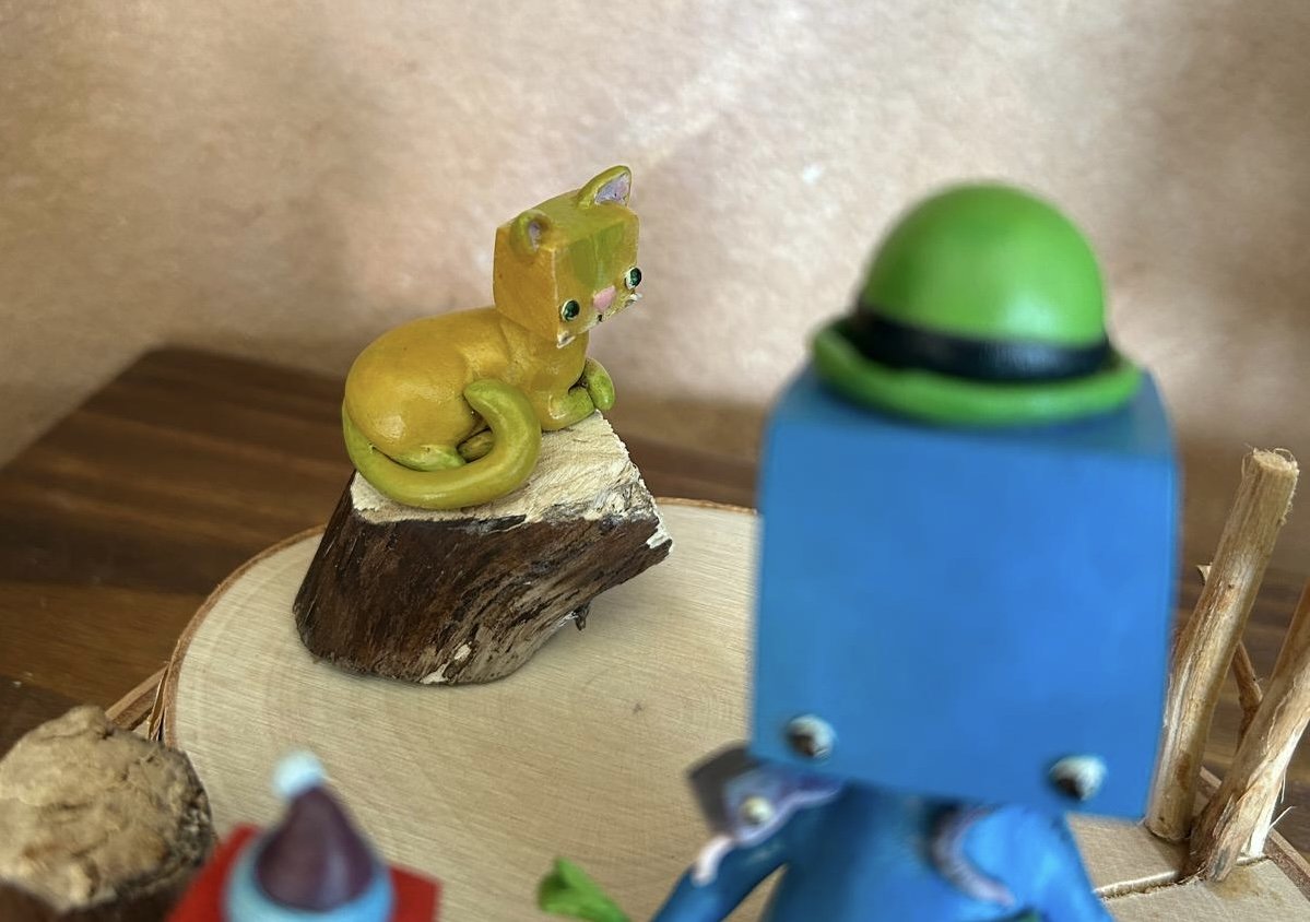 A decorative scene with a small yellow cat figurine with large green eyes sitting on a piece of wood, and a blue box-head with a green hat in the foreground. The background features a beige wall and a wooden surface.