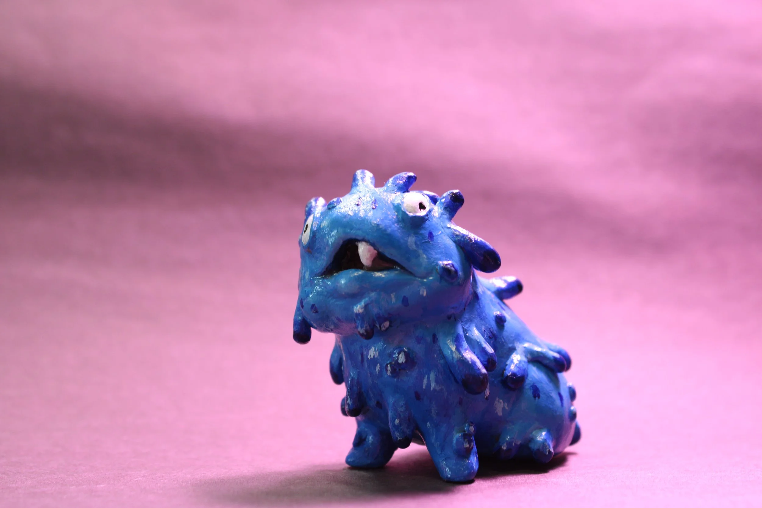 A small, blue, dinosaur-like figurine with multiple rounded protrusions on its back and head, standing on a pink surface.