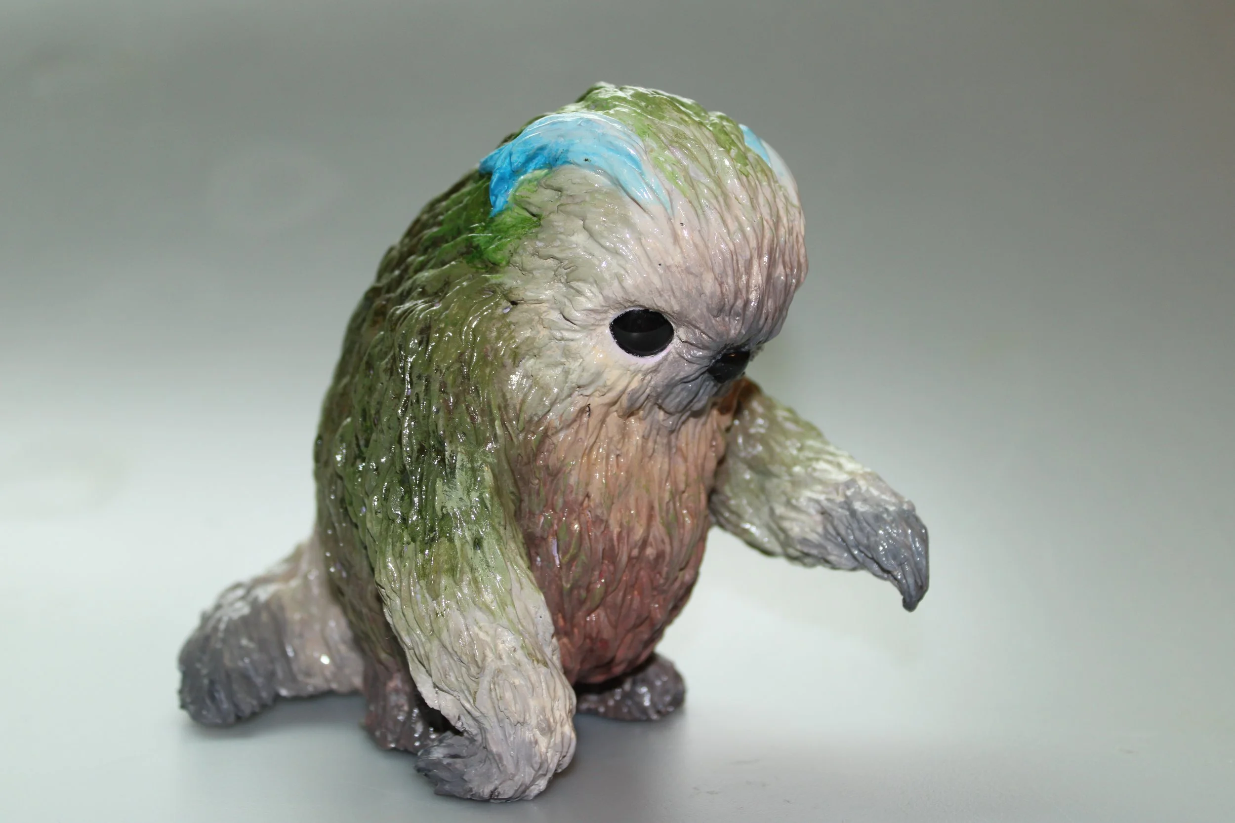 A sculpture resembling a cute, chubby creature with a rounded body, large dark eyes, and long arms. Its surface is textured with a mix of green, beige, and pinkish colors.