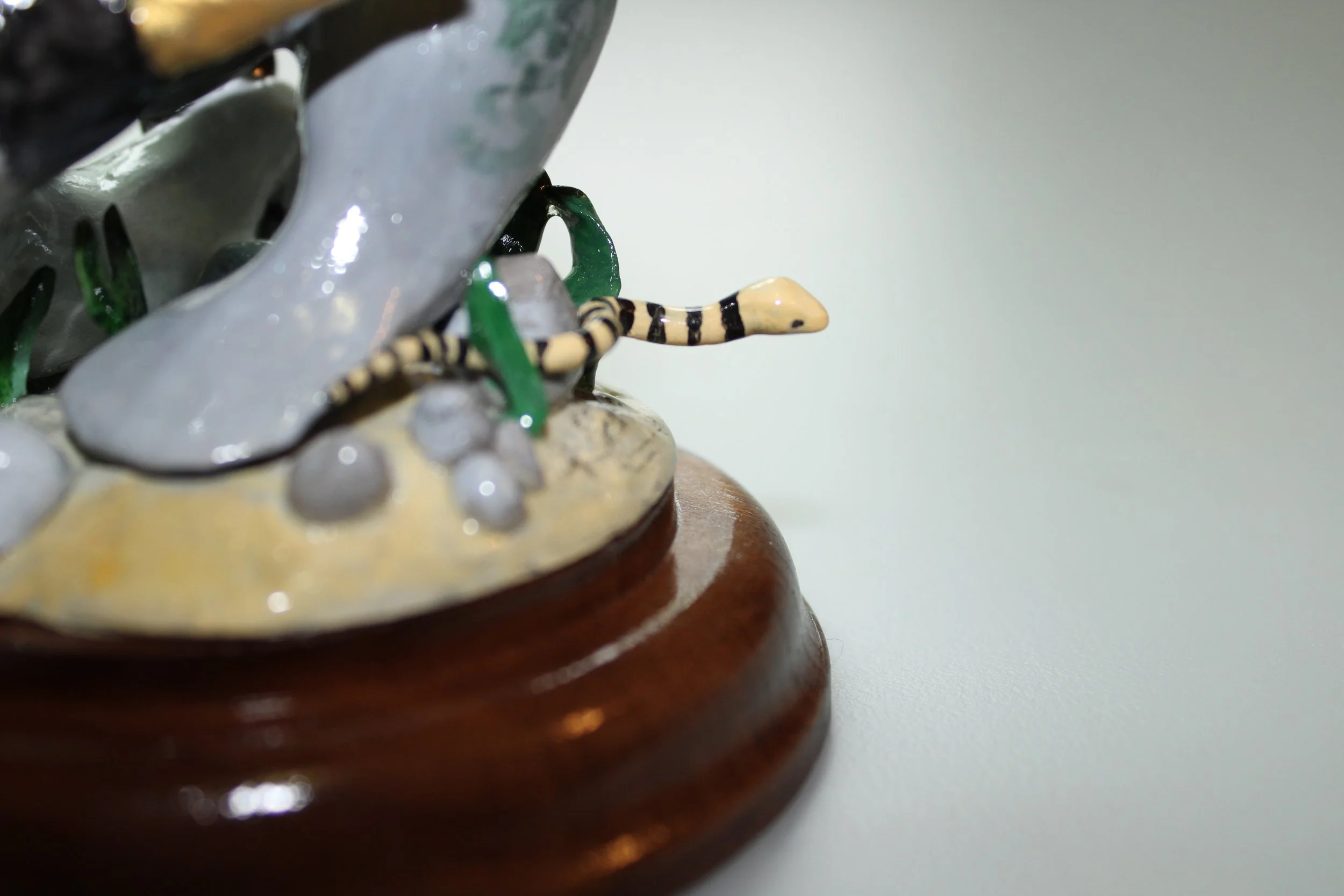 Close-up of a clay sculpture featuring a yellow and black striped snake, with green leaves and gray rocks, on a brown wooden base.