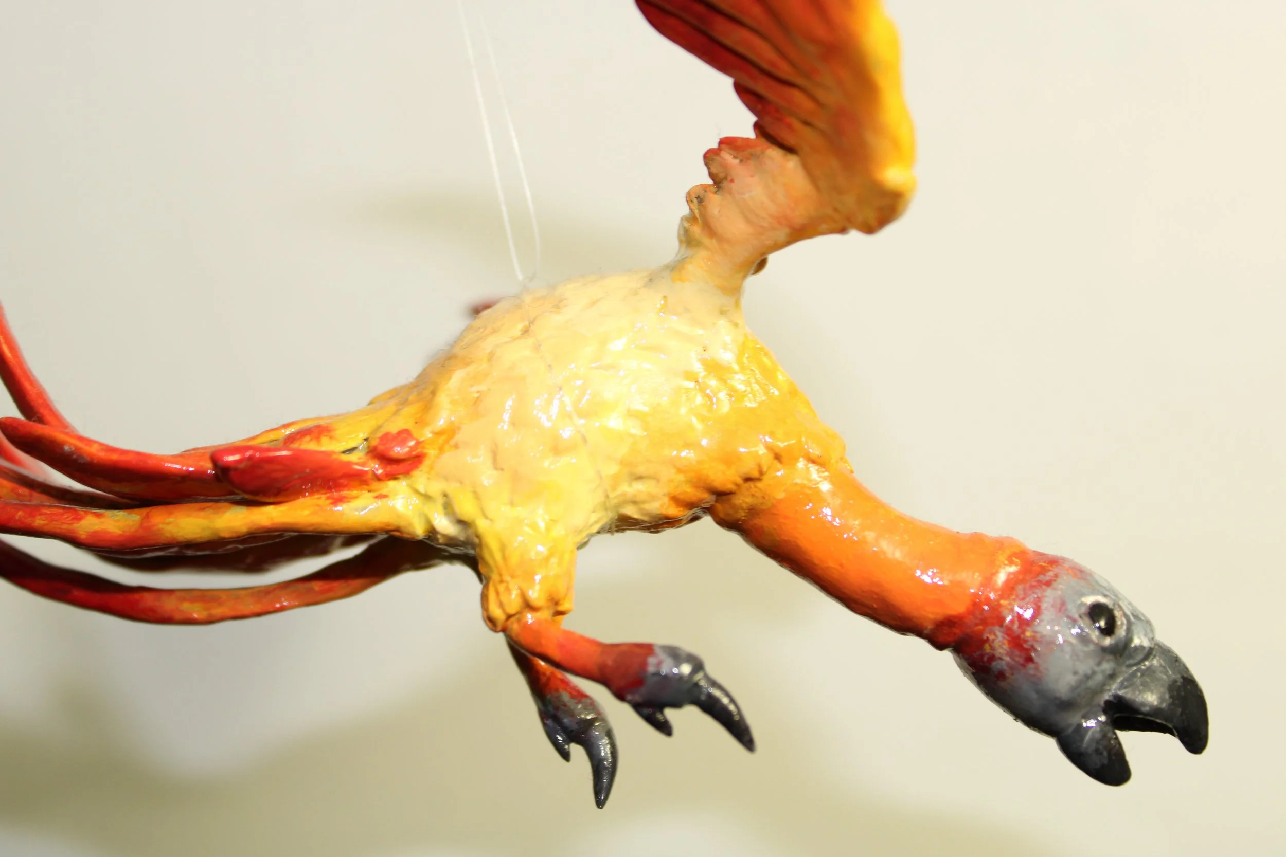 A sculpture of a bird with bright orange and yellow feathers, black claws, and a black beak, hanging against a plain background.