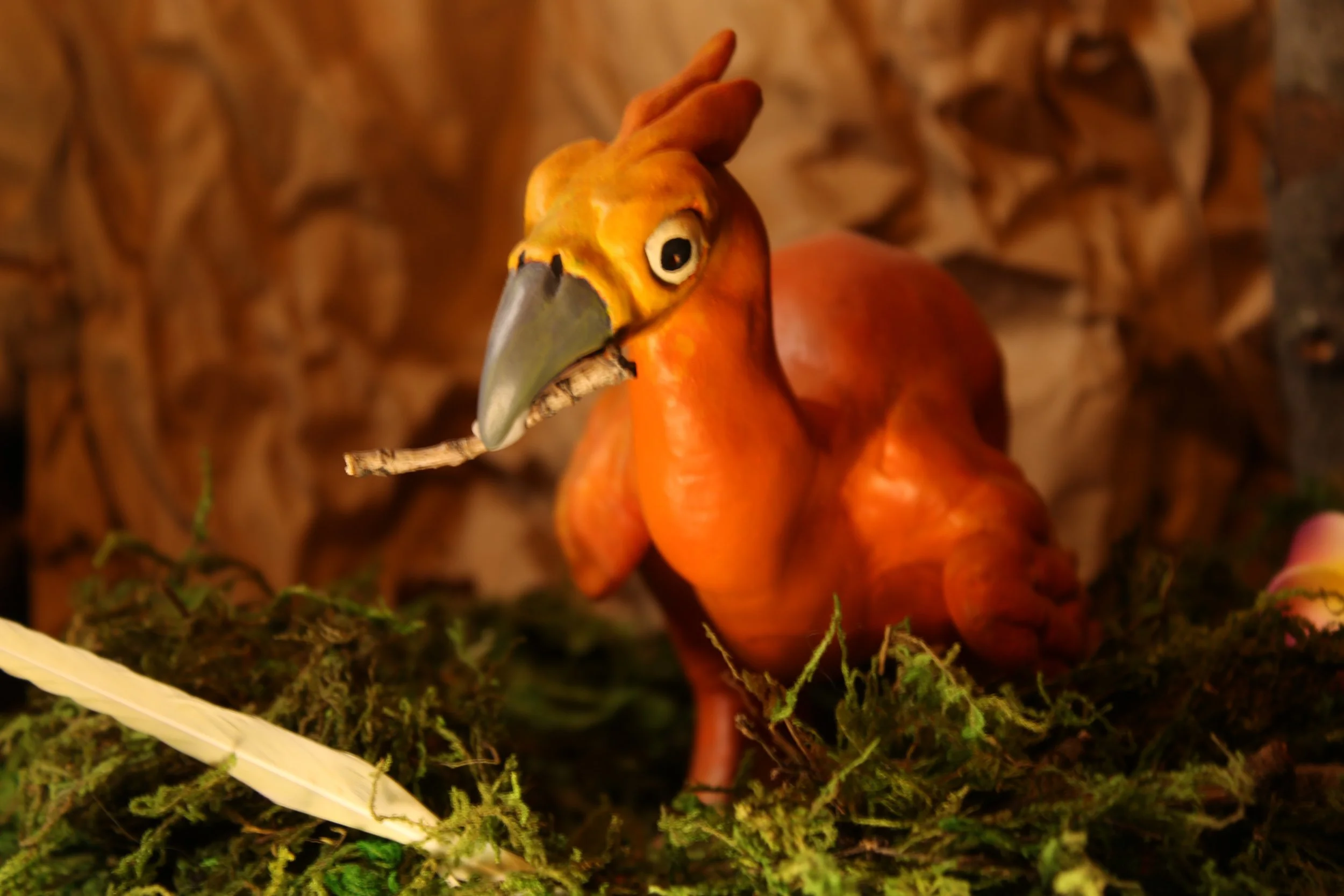 A crafted figurine of a toucan bird with a large beak, sitting on moss and holding a small stick in its mouth, in a naturalistic setting.