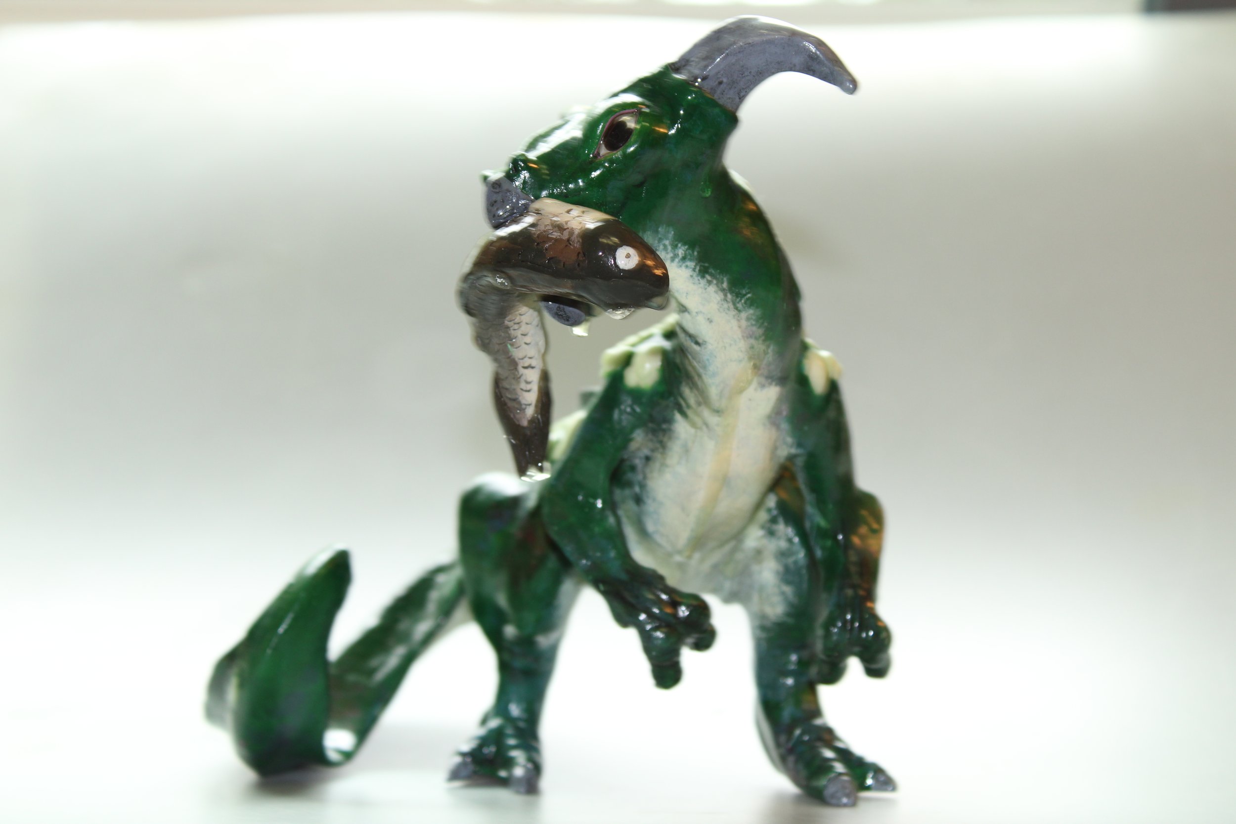 A clay figurine of a green and white dragon holding a fish in its mouth, with a gray horn on its head and a tail curled downward.