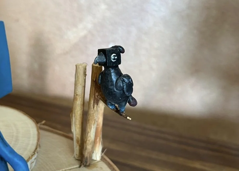 Small black figurine of a bird with white eyes and a beak, perched on two sticks, against a plain wall.