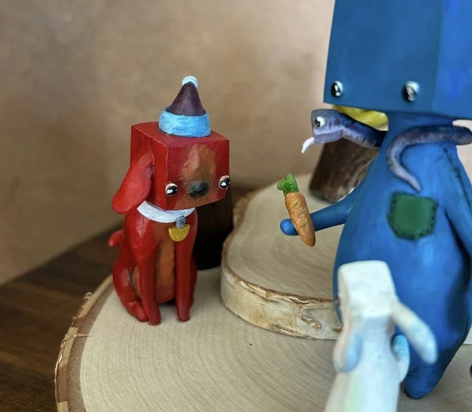 Colorful figurines of a dog with a party hat and a person in blue holding a carrot, set on a wooden surface.