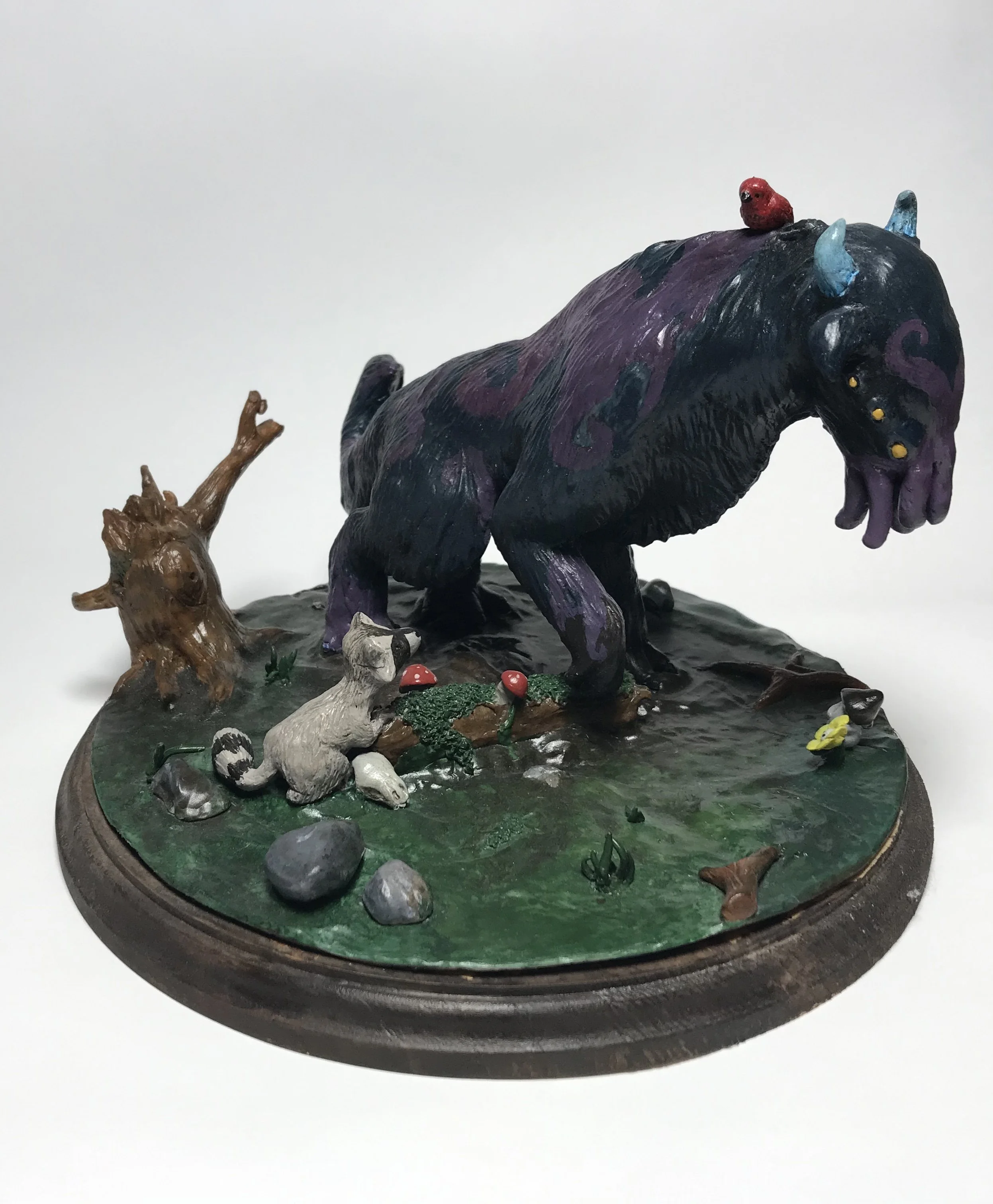 A colorful sculpture of a whimsical creature resembling a bear or monster with horns, sitting on a base with miniature elements like a raccoon, mushrooms, a squirrel, and tree branches, set on a white background.
