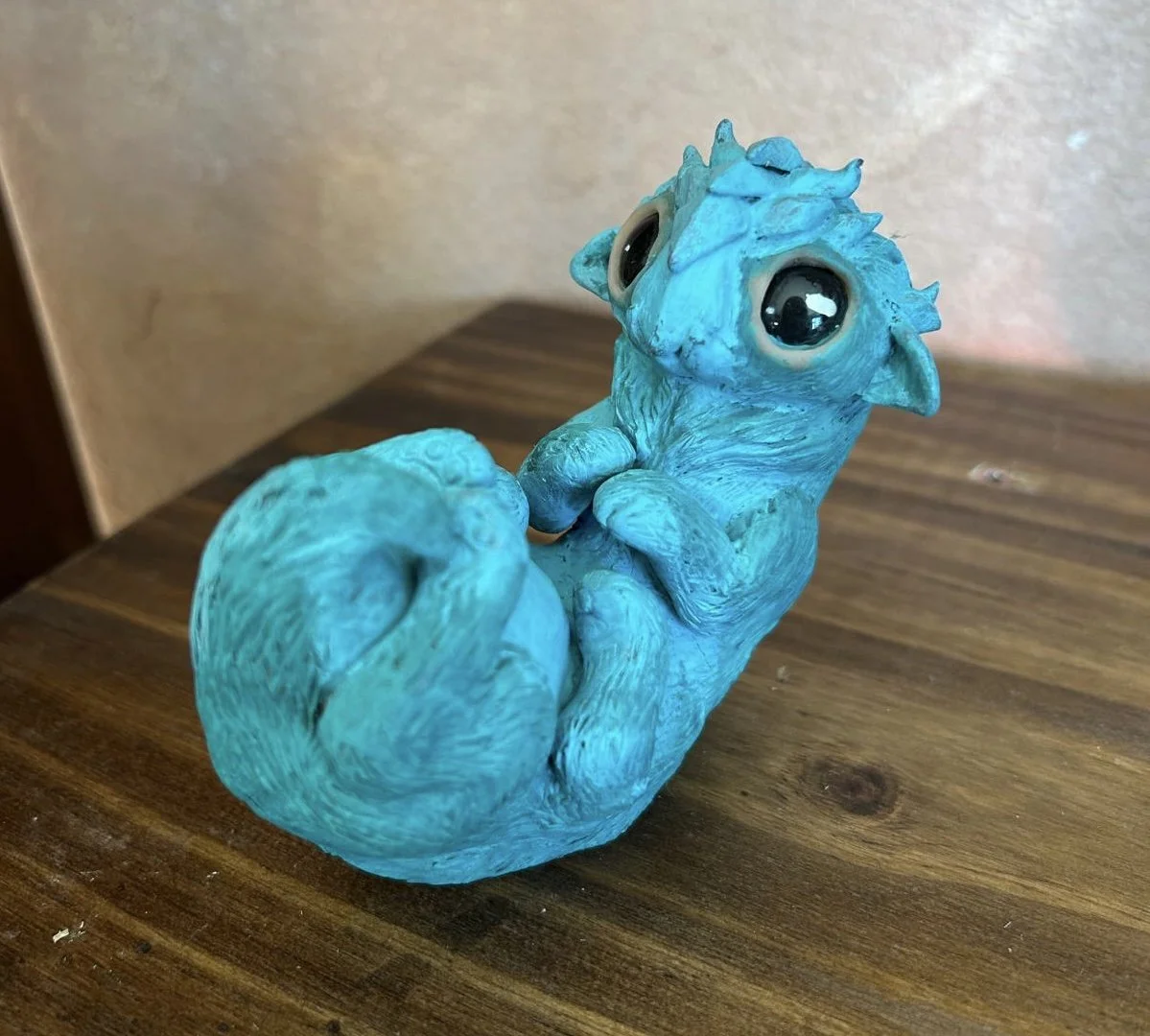 A blue, cartoonish dragon figurine with large black eyes, sitting on a wooden surface, facing upward.