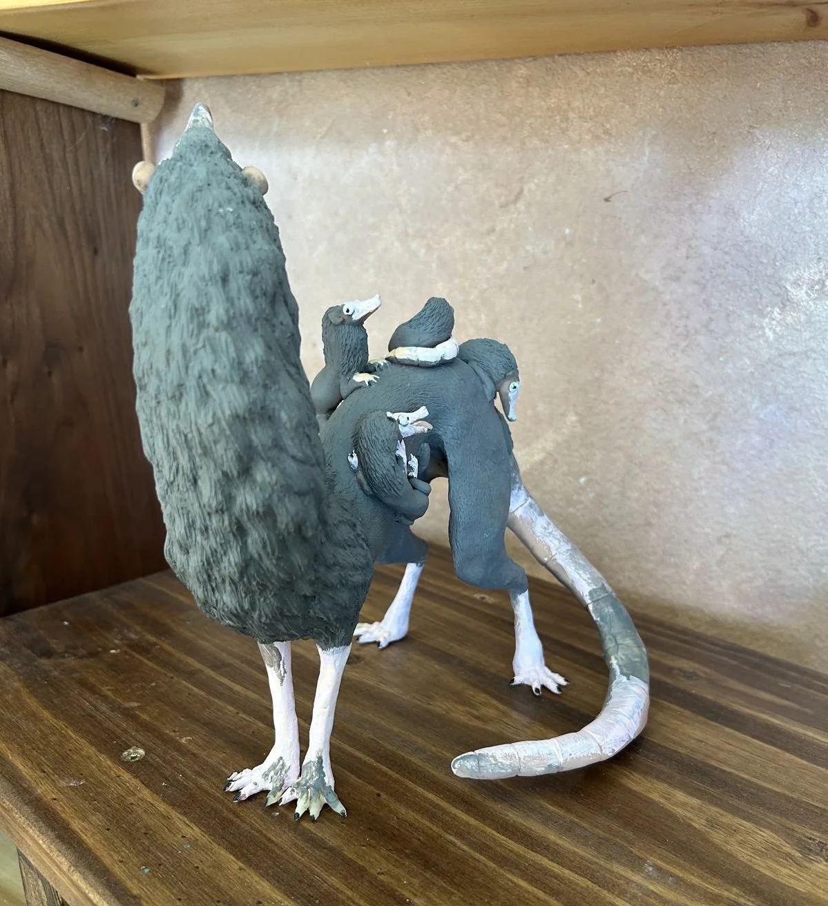 A detailed figurine of a strange long necked opossum, filled with smaller versions of itself on its back, positioned on a wooden surface against a beige background.