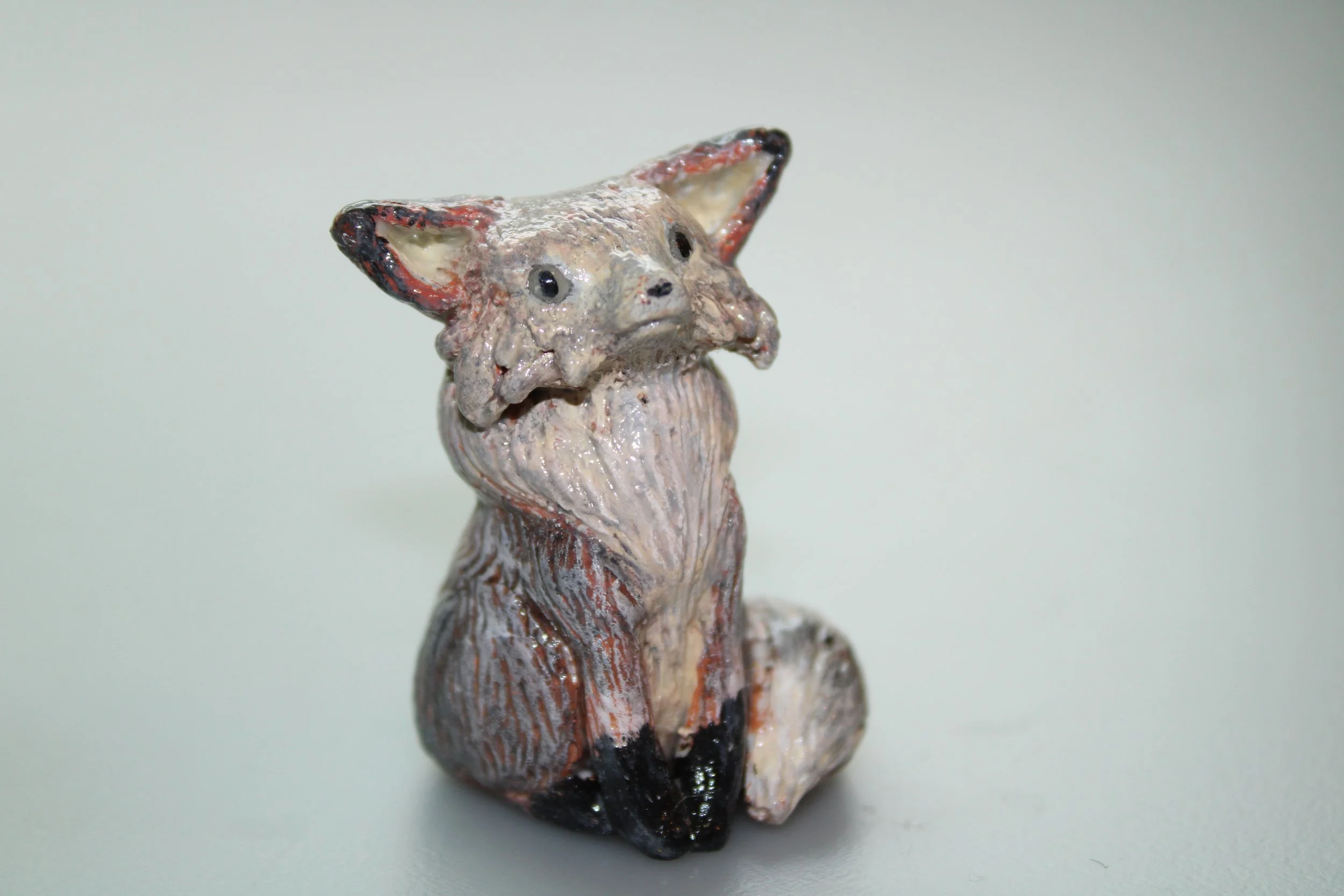 Clay sculpture of a fox with exaggerated ears and, sitting on a plain light-colored surface.