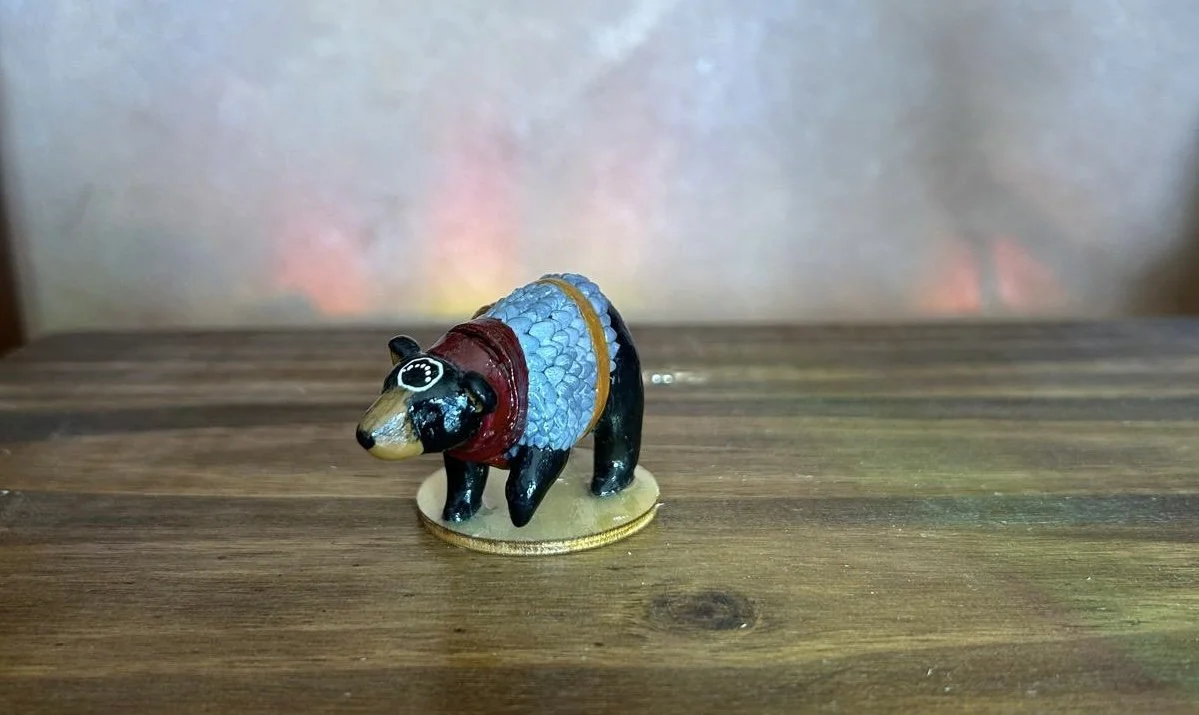 Small figurine of a bear wearing a red scarf and scaled armor, standing on a round base on a wooden surface.
