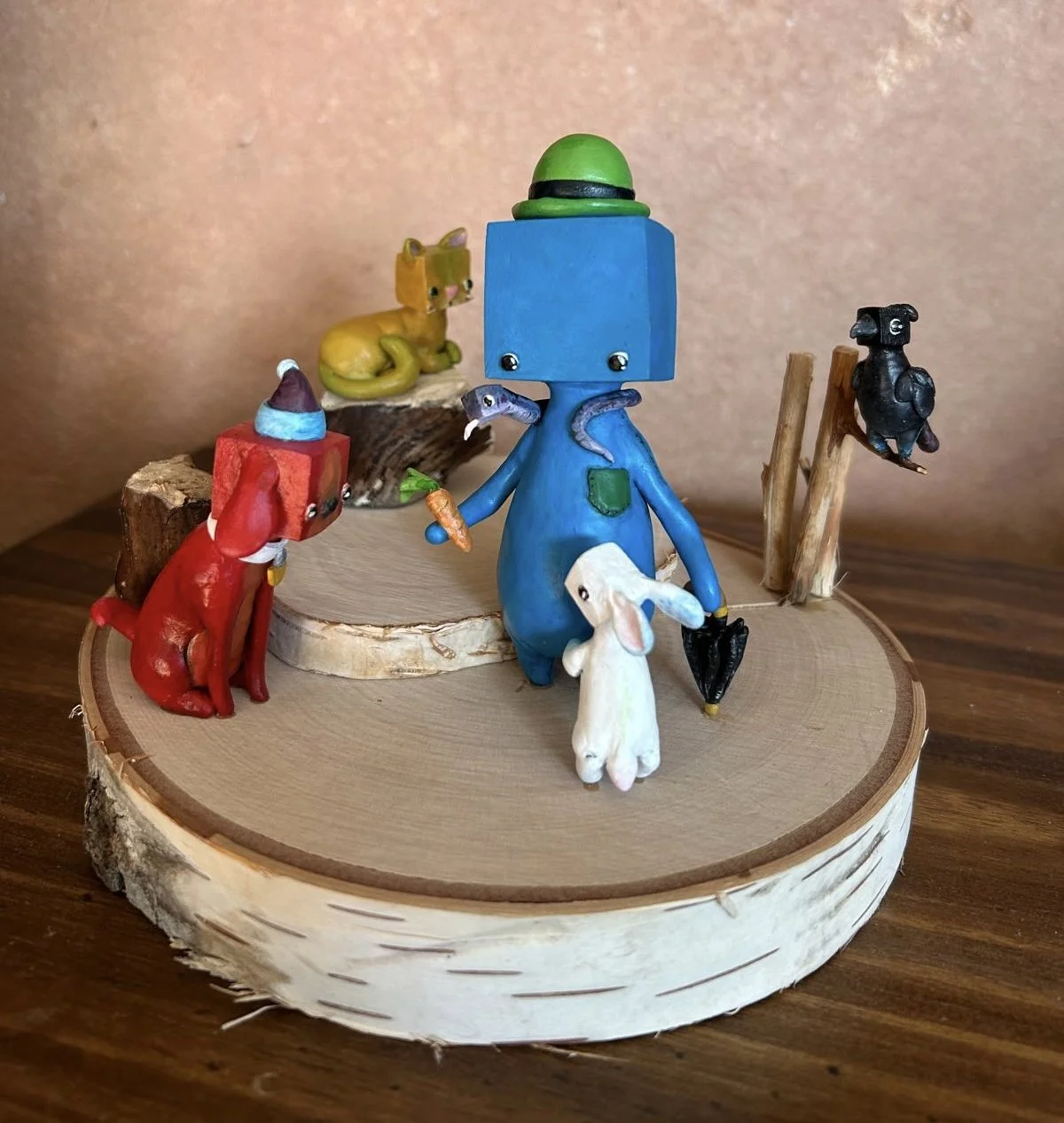 Miniature animal figurines arranged on a wooden round display, including a blue cube headed person holding a carrot and umbrella, a red dog with a Santa hat, a white rabbit, a yellow cat, a black bird, and a small wooden log background.