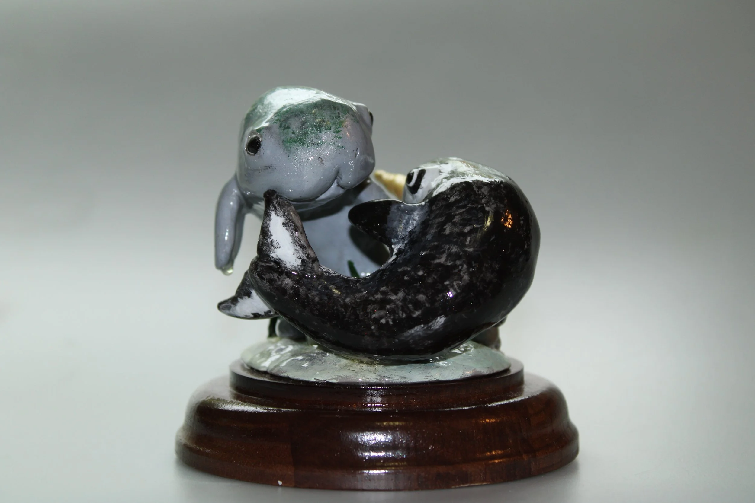 A clay figurine sculpture of two animals, a narwhal and a manatee, intertwined on a round wooden base.
