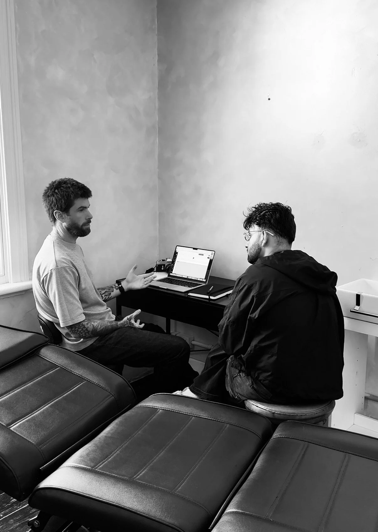 Two men having a conversation in an office. The man on the left is gesturing with his hands, seated at a desk with a laptop, notebook, and pen. The man on the right, wearing glasses and a dark jacket, is sitting on a stool facing the first man. The office has plain walls, a window with blinds, and a row of benches in the foreground.
