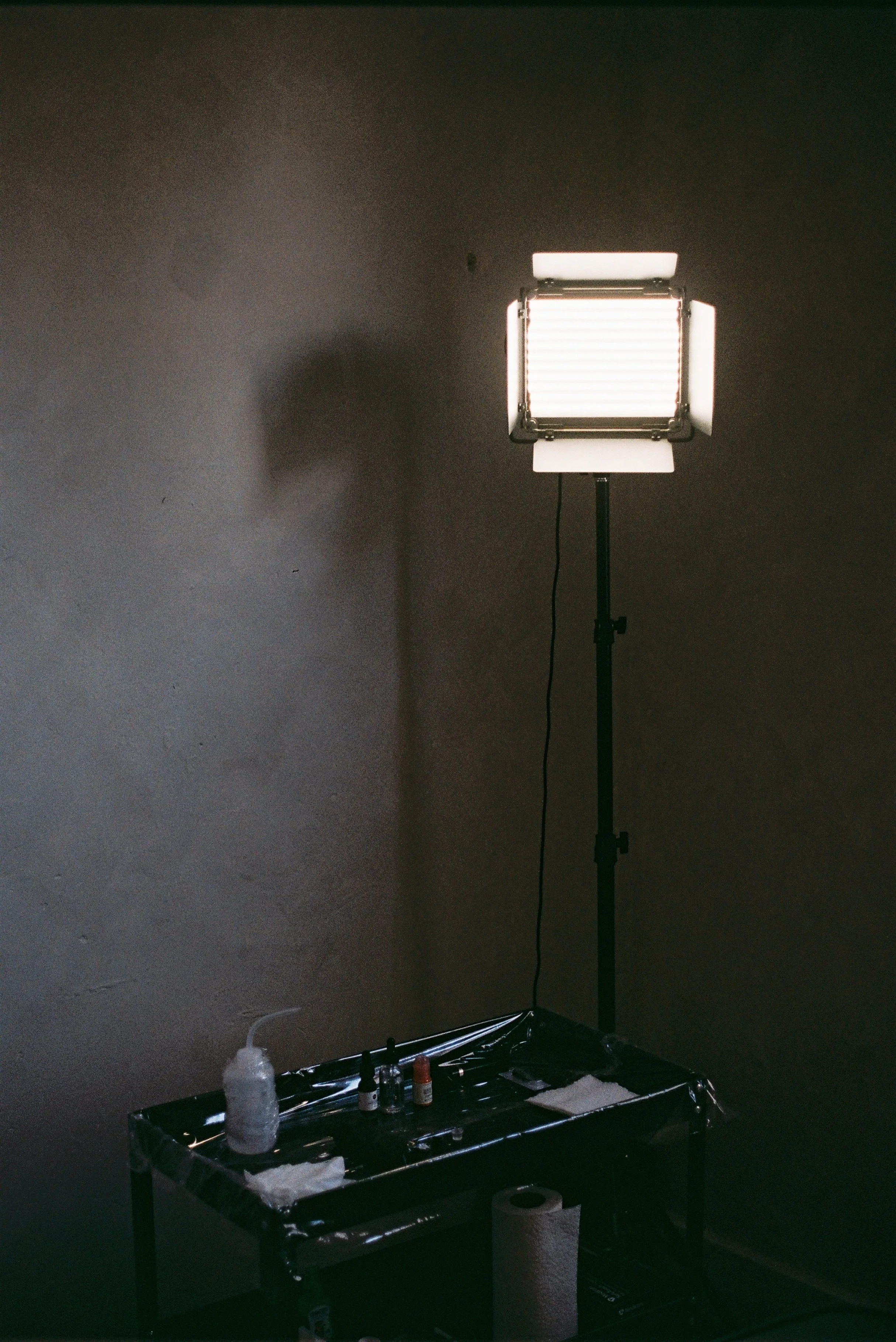 A photography lighting setup with a large bright LED light on a stand in a dark room, with a tray of supplies, paper towels, and spray bottles on a table underneath.