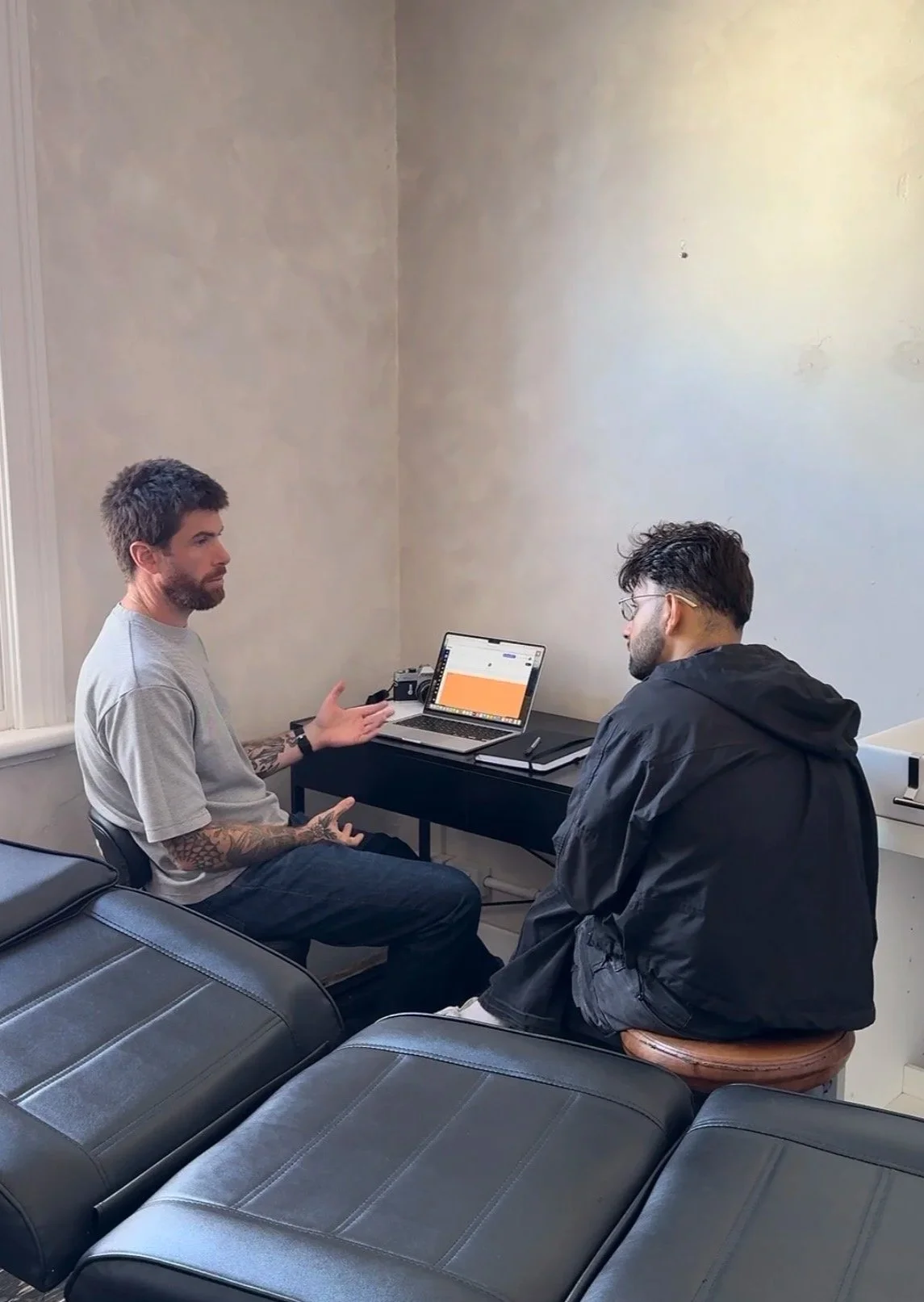 Two men are having a conversation in a room with minimal decor. One man with tattoos and short hair is gesturing with his hands, sitting next to a laptop on a black table. The other man, with glasses and darker hair, is sitting on a round stool facing him. There are black leather couches in the foreground.